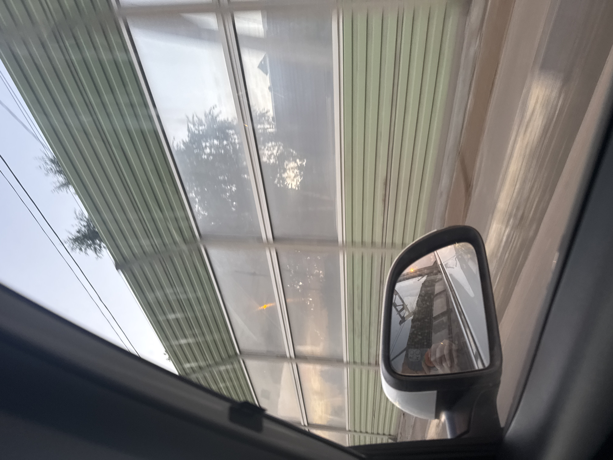 The image shows a view from the passenger side of a car moving along a road, likely in Mstikhino, Russia. A sound barrier, constructed of green corrugated metal panels and translucent inserts, runs parallel to the road. The inserts reflect the sky and trees, and a single yellow light is visible through one of them. The car's side mirror is also visible, reflecting a person holding a phone and the road ahead, which appears wet. The sky is overcast with hints of sunset.