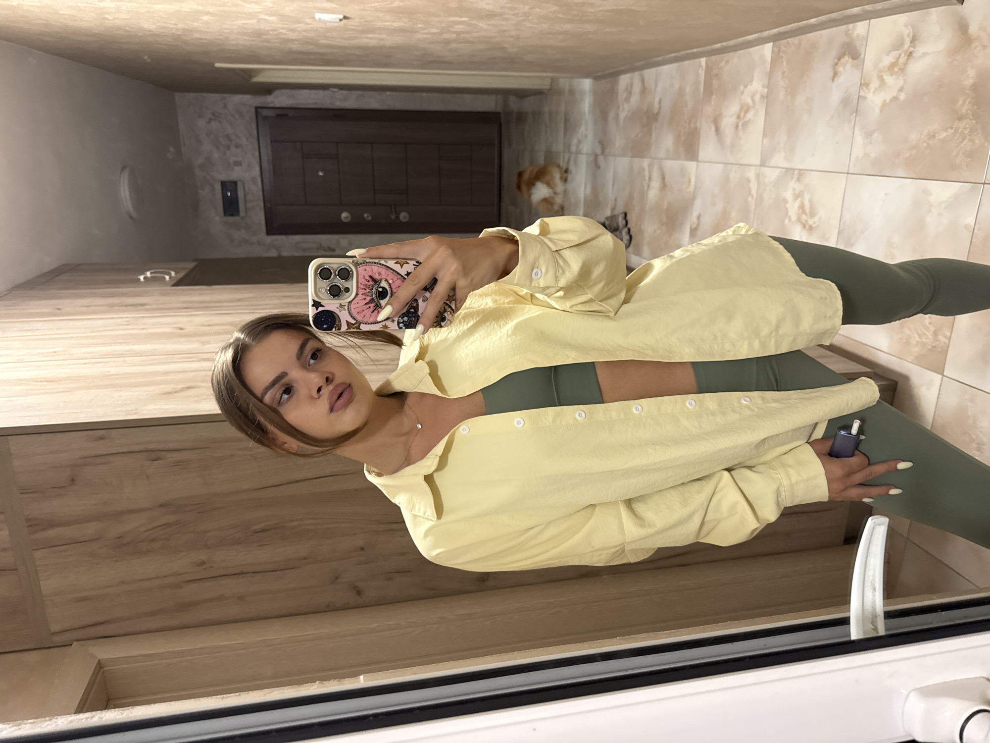 A young woman with brown hair and light skin is taking a selfie in a hallway with light brown walls and tiled flooring. She is wearing a yellow button-down shirt over a green sports bra and green leggings. The woman is holding a phone with a pink and white case adorned with an eye design. In the background, a small dog with light brown and white fur is lying on the floor. The hallway appears to be in a residential building.