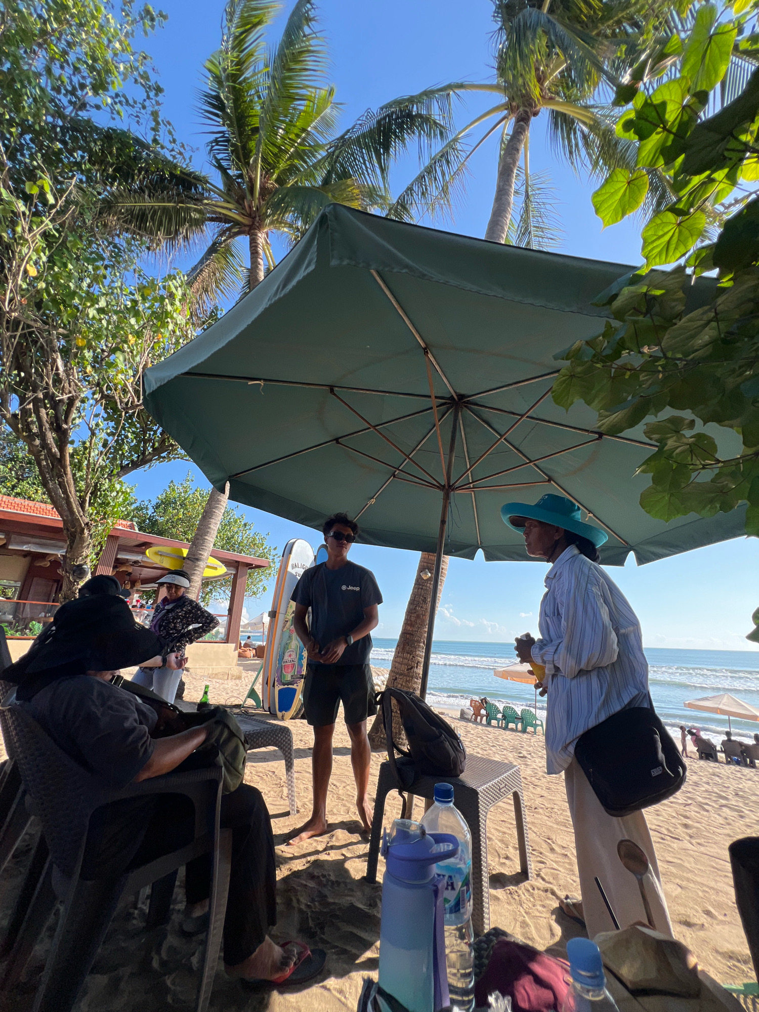 The image depicts a group of people on a sandy beach in Kuta, Indonesia, near a large green umbrella. In the foreground, a person wearing a wide-brimmed black hat and dark clothing is seated in a black plastic chair. To their right, another person in a dark t-shirt with a "Jeep" logo and dark shorts stands with sunglasses. Two surfboards are propped up behind them. To the right of the standing person, a woman wearing a light blue hat and a striped long-sleeved shirt is holding a yellow bottle and looking to the left. A black shoulder bag is slung across her body. On a table in front of her are water bottles, one blue and one clear with blue lettering. The background shows the ocean, palm trees, and other beach umbrellas. A building with a red roof is visible on the left. The lighting suggests it is a sunny day.