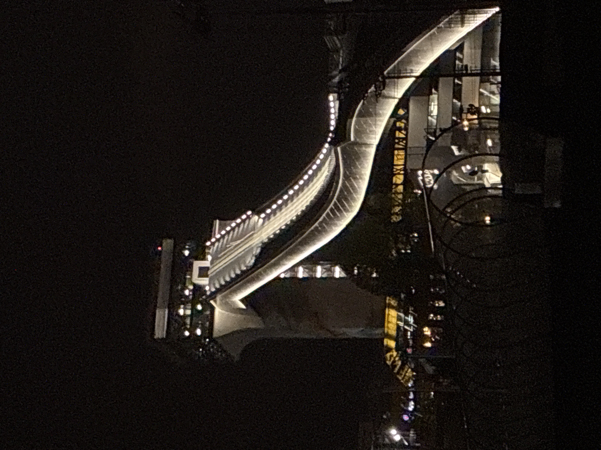 The image shows a bridge illuminated at night in Moscow, Russia. The bridge is a modern design with a curved walkway and decorative lighting. Reflecting below is what appears to be a body of water and its reflection is visible in the water. Other buildings are visible to the right of the bridge and its reflection is in the body of water. The background is dark and obscures details.