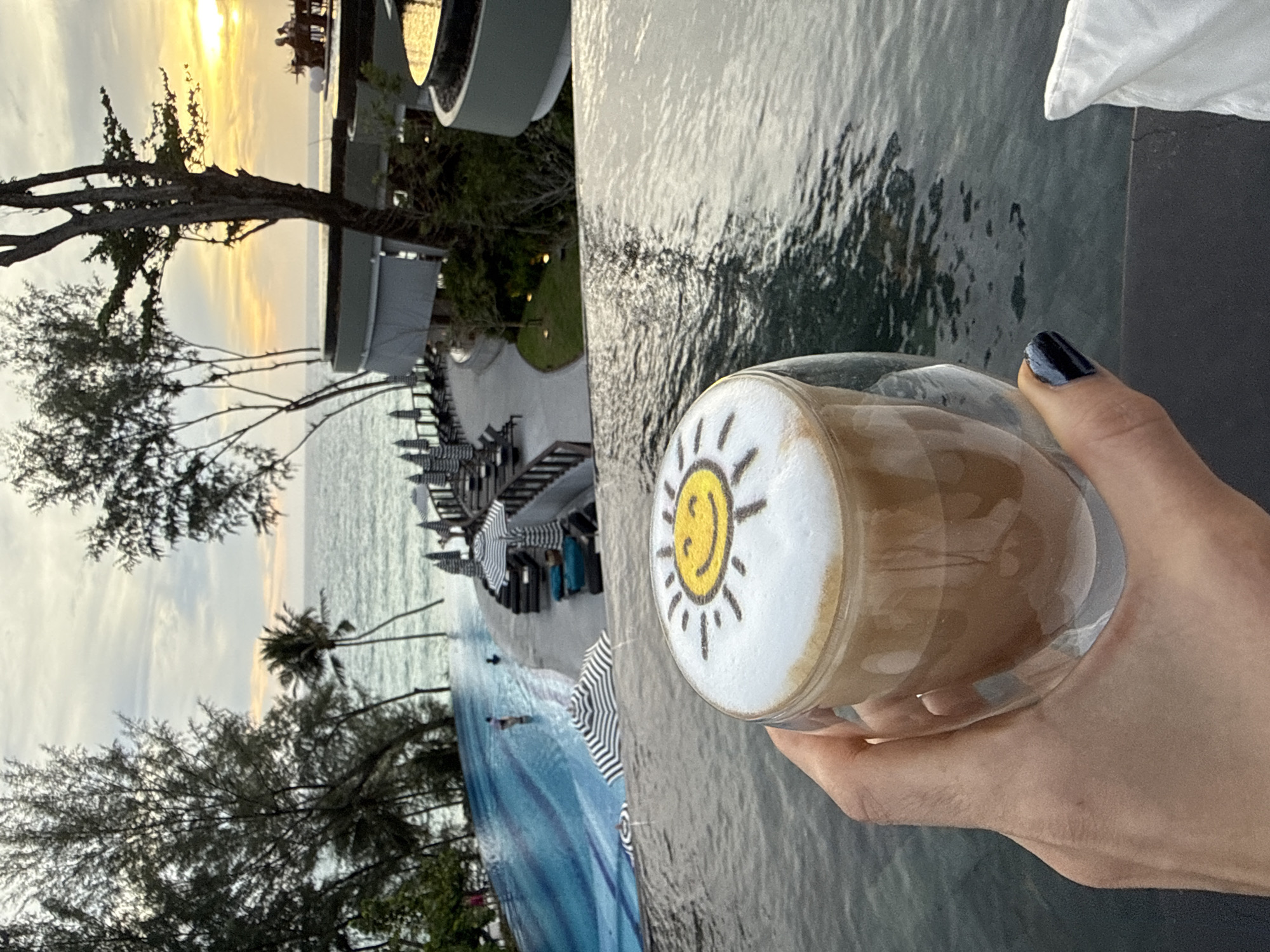A hand with dark blue and black nail polish holds a clear glass of latte with a sun design on the foam. The latte is in the foreground, with a blurry infinity pool and a beach resort in the background. The sky is overcast with some golden light from the sunset peeking through. There are palm trees and other vegetation, as well as lounge chairs and umbrellas by the pool. Two people are visible in the pool. The location is Sakhu, Thailand.