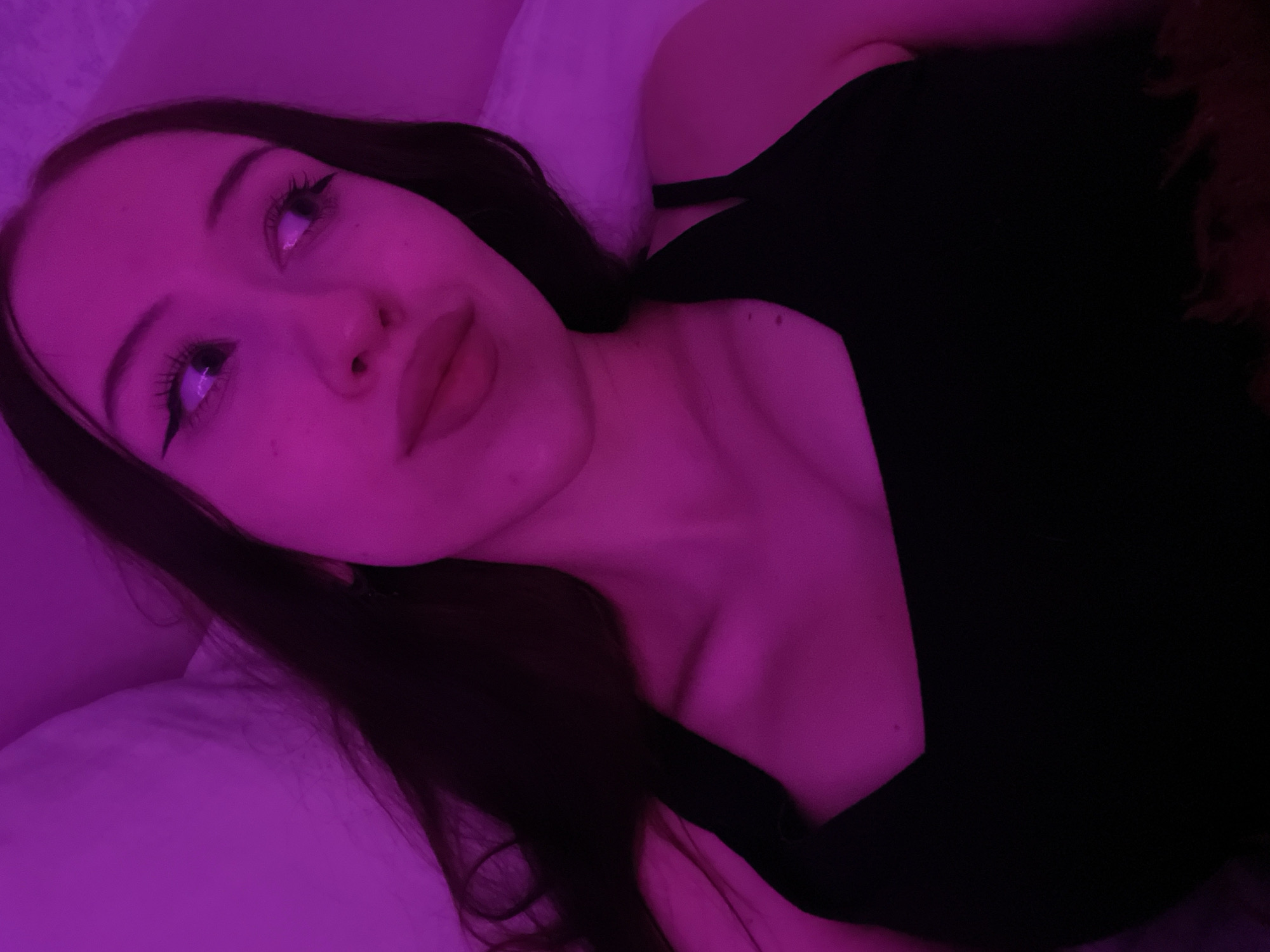 A young woman with long, dark hair and makeup, including winged eyeliner, is shown in a close-up shot. She is wearing a black tank top and lying down, with a pink light illuminating her face and upper body. The background appears to be a bed with pink bedding. There is no information in the image that indicates this scene is located in Votkinsk, Russia.