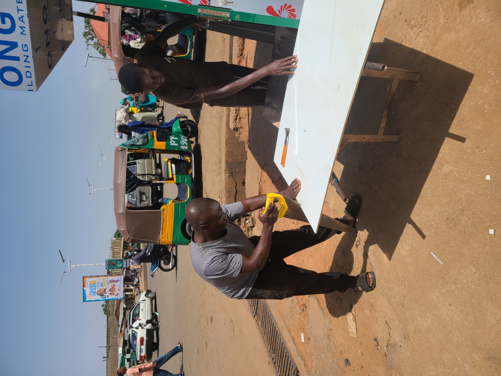 Two men are cutting a white board on a city street. One man, dressed in a grey t-shirt and dark pants, is sawing the board with a small yellow hand saw. The second man, wearing a brown shirt and dark pants, is steadying the board. The board sits on a makeshift table comprised of metal legs. An auto rickshaw is visible behind the men, and there are also buildings, vehicles, and pedestrians in the background.
