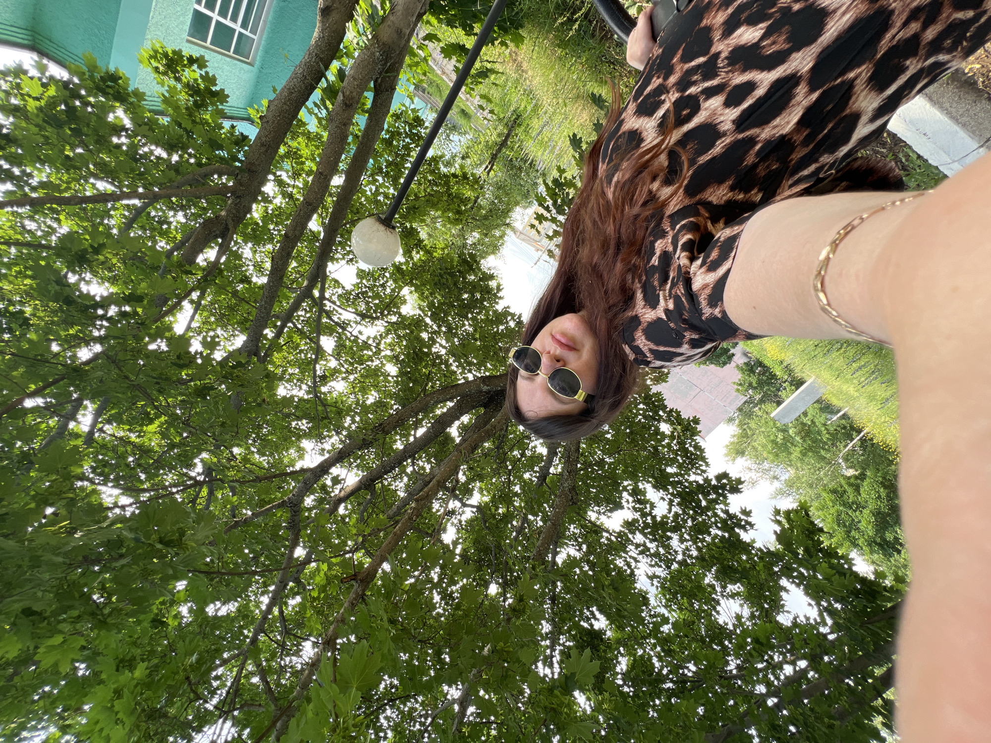In Noviy, Russia, a woman with long, auburn hair and sunglasses is captured in a low-angle, selfie-style shot. She is wearing a leopard-print dress and a gold bracelet is visible on her arm. The background features lush green foliage from a large tree overhead, with a bright sky peeking through the leaves. To the right, a turquoise building with a window is partially visible, and a lamppost with a spherical light fixture stands nearby. The scene appears to be outdoors, possibly in a park or residential area, on a bright, sunny day.