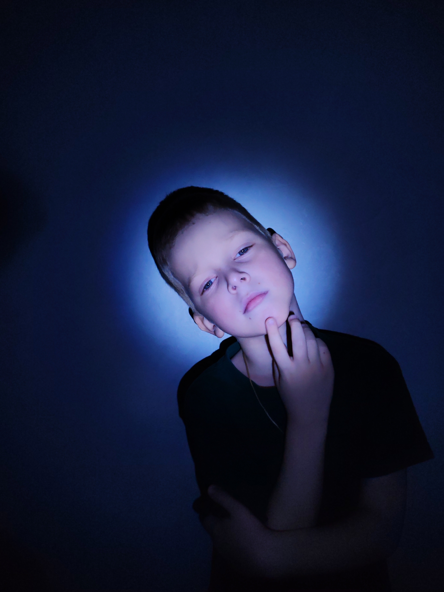This portrait features a young boy, likely in his pre-teen years, centrally positioned against a dark background. A prominent circular spotlight of bright blue light illuminates the area directly behind and around his head, creating a halo effect that fades into deep blue and black tones towards the edges of the frame.
The boy has short, light-colored hair and light blue eyes, which are directed straight towards the viewer. He is wearing a dark, possibly black or dark green, short-sleeved t-shirt, and a thin gold-colored chain or necklace is visible around his neck. His right hand is raised, with his fingers gently touching his chin and jawline, suggesting a thoughtful or posed expression. His left arm is bent, with the forearm resting across his body, partially obscured by his shirt. His facial expression is subtle, appearing neutral, slightly serious, or pensive.
The setting is an indoor space, indicated by the plain, untextured background and artificial lighting. There are no specific environmental cues to suggest a particular room or time of day, as the lighting is entirely controlled and stylized. No activity beyond posing for the photograph is depicted. There is no visible text in the image. The provided location context for this image is Chaikovsky, Russia.