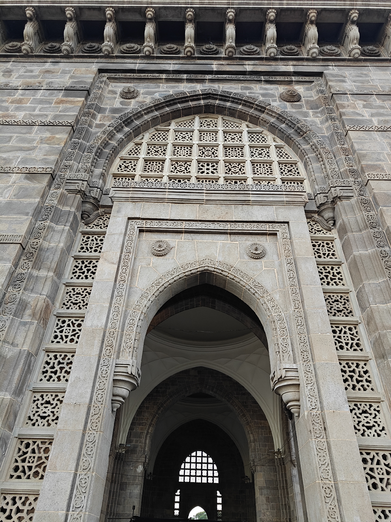The image shows a close-up, low-angle view of the Gateway of India in Mumbai, India. The architectural details are prominent, including the intricate latticework, arches, and decorative carvings on the stone structure. The view is focused on the central arch and the layers of construction leading upward. A glimpse of the sky and surrounding area is visible through the arches in the background. The building appears weathered and historic.