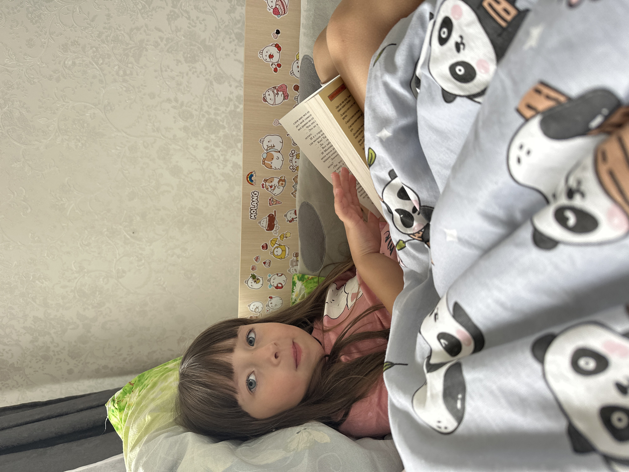 In the city of Chaikovsky, Russia, a young girl with long brown hair and blue eyes is lying in bed, propped up against a pillow, reading a book. She is wearing a pink shirt with a white panda design. The bed is covered with a light blue duvet featuring a pattern of panda faces. Behind her, the headboard of the bed is decorated with various stickers of cartoon characters. The background wall has a textured floral wallpaper. The scene appears to be indoors, likely a bedroom, with natural light coming from the left.