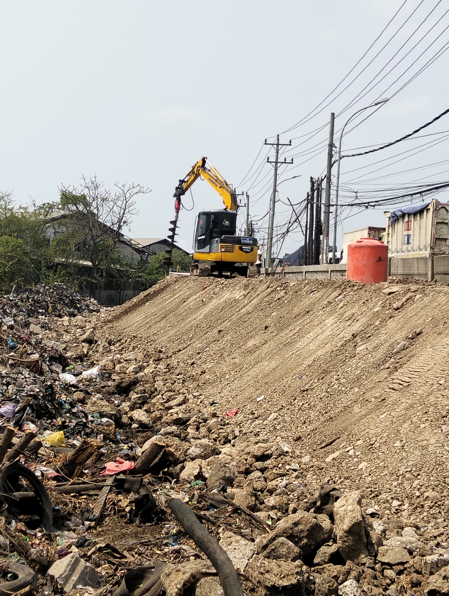 The image depicts a construction site in Semarang, Indonesia, characterized by a significant amount of debris and heavy machinery. In the foreground, a large pile of garbage and rubble, including tires, plastic, and various discarded materials, is visible. To the right, a graded dirt mound, likely prepared for construction, dominates the mid-ground. A yellow Komatsu excavator with a drilling attachment is positioned on top of this mound, actively engaged in drilling. In the background, several utility poles with a complex network of electrical wires crisscross the sky. A large orange water tank stands near a concrete barrier, with a truck visible behind it. Buildings and trees are also present in the distance, suggesting an urban or semi-urban environment. The overall impression is one of ongoing development or renovation amidst a challenging environmental context.