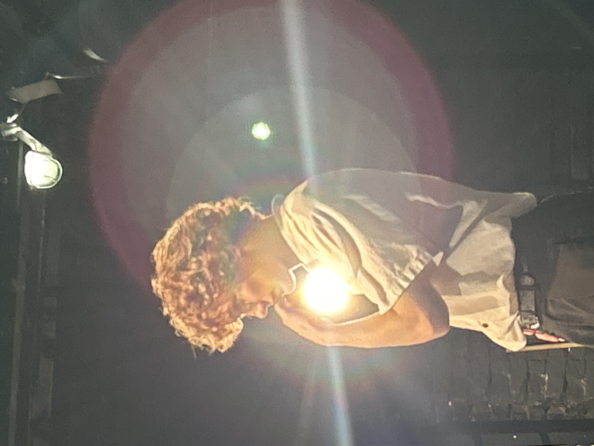 The image shows a person with curly, light brown hair, leaning forward with their head down. They are wearing a light-colored, possibly white, short-sleeved shirt. The person is holding a bright light source, perhaps a flashlight or a bare lightbulb, close to their face. The light is intense and creates a significant glare and lens flare, obscuring some of the details of the scene.
The background is dark and out of focus, suggesting an indoor setting or nighttime outdoors. There is a visible light fixture on the left side of the frame, emitting a soft glow. The overall impression is one of concentration or introspection, with the bright light drawing attention to the person's face and hands. The presence of lens flare suggests the photo was taken directly towards a light source, possibly by a camera phone or digital camera with a less sophisticated lens. There is no visible text in the image. The lighting conditions are dramatic and moody.