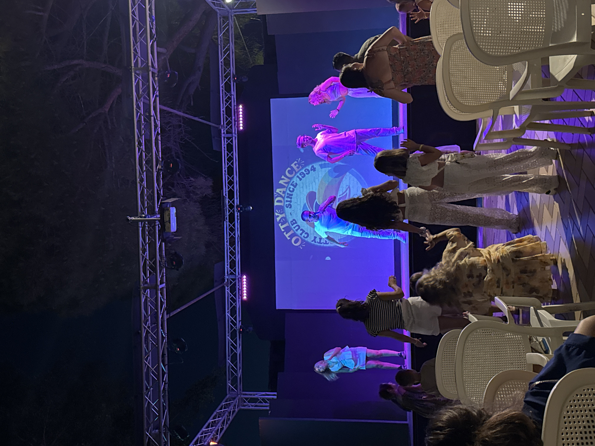 This image captures an outdoor evening event at the "OTTY DANCE CLUB SINCE 1994" in Isola delle Femmine, Italy. A stage is set up with a large screen displaying the club's logo, illuminated by purple and pink stage lights. Five dancers, three men and two women, are performing on the stage, some in casual wear and others in more active attire. A crowd of spectators, including adults and children, are seated in white chairs in the foreground, facing the stage and observing the performance. The lighting suggests it is nighttime, with artificial lights from the stage and surrounding area illuminating the scene.
