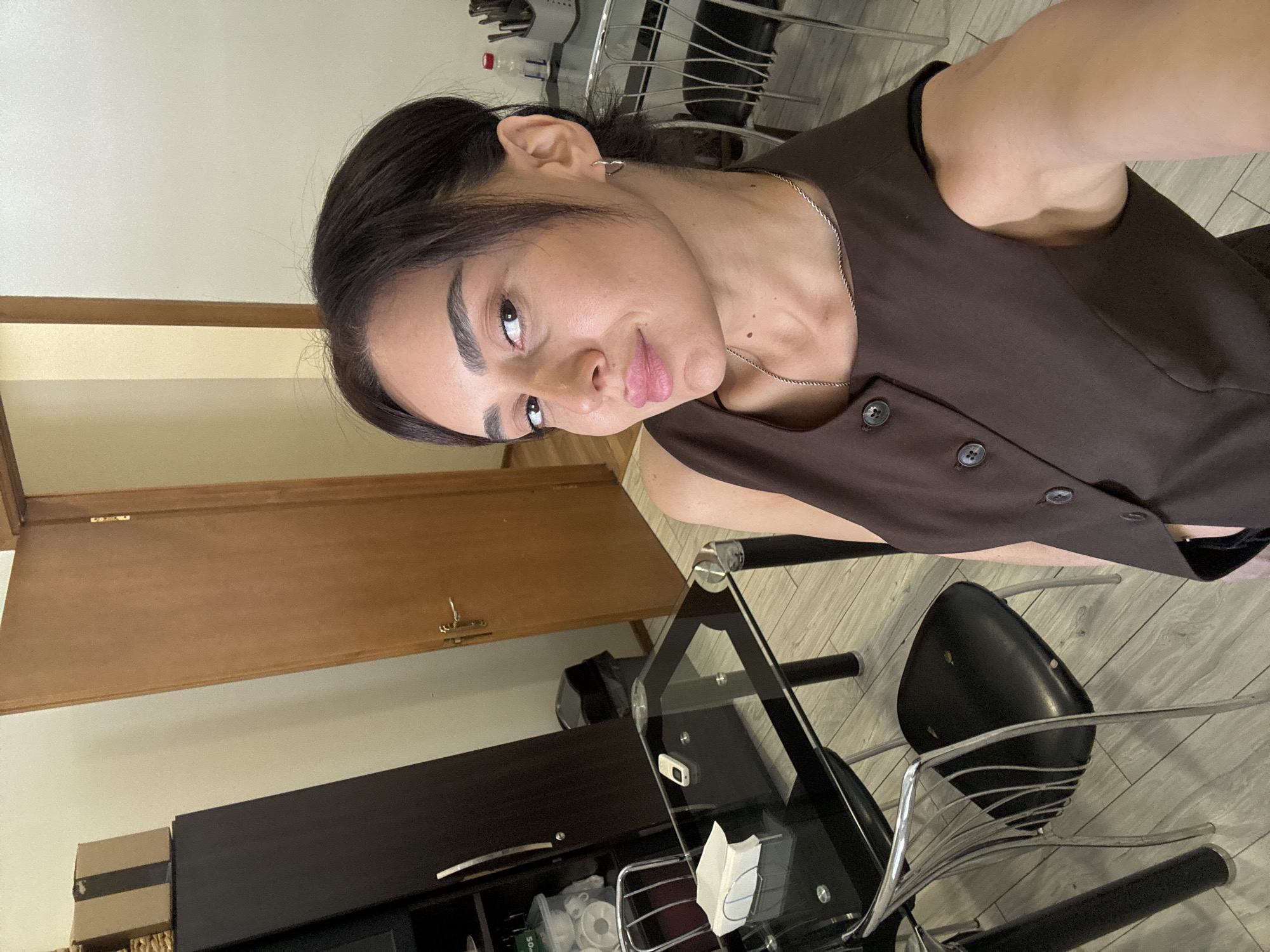 A young woman of Middle Eastern descent, with dark hair pulled back and significant makeup, is taking a selfie in a room that appears to be a dining area. She is wearing a brown sleeveless vest with two visible buttons. The room has light-colored wood-plank flooring and a light-colored wall. To her left is a dark brown tall cabinet topped with a cardboard box. In front of the cabinet is a black glass-top dining table with metal legs and chairs. On the table are a white napkin holder and a small electronic device. A trash can is visible behind the table. To her right, a wooden door with a brass handle and hinges is slightly ajar. In the background, to the right of the door, is another table with metal chairs, a container of utensils, and a plastic bottle. The lighting in the room is artificial and appears to be coming from overhead. The woman is looking directly at the camera with a slight smile. There is no indication this image was taken in Tbilisi, Georgia.