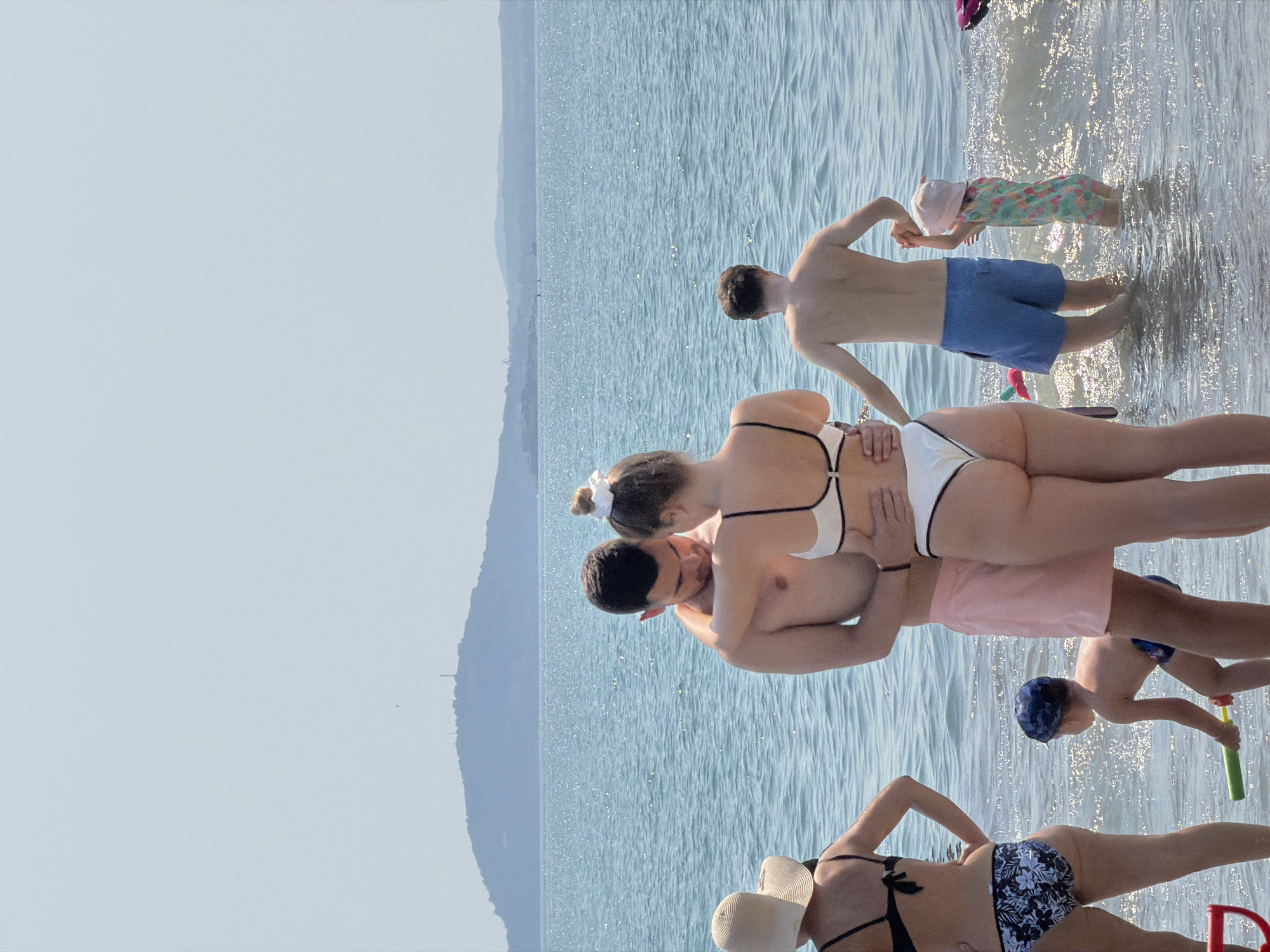 The image depicts a sun-drenched beach scene in Burgas, Bulgaria, with several people enjoying the water on a clear, bright day. The sun glints off the surface of the calm, blue sea, creating a shimmering effect.
In the foreground, two young adults are embracing in the shallow water. The woman is wearing a white bikini with black trim, and the man is shirtless. Behind them, a young boy in blue swim trunks and a blue cap is playing with a green water gun, appearing to be in motion. Further back, a man in blue swim trunks stands in the water, facing away from the camera. A toddler, wearing a floral-patterned outfit and a pink cap, is also in the water near the man, seemingly being held by him or walking near him.
In the lower part of the image, a woman in a black and white floral bikini and a straw hat is seated in the shallow water, facing away. Another person is partially visible behind her, wearing a black bikini.
The background shows a hazy shoreline with hills or mountains in the distance, consistent with the landscape of Burgas. The overall atmosphere is one of leisure and enjoyment on a warm summer day at the beach. There is no visible text in the image.