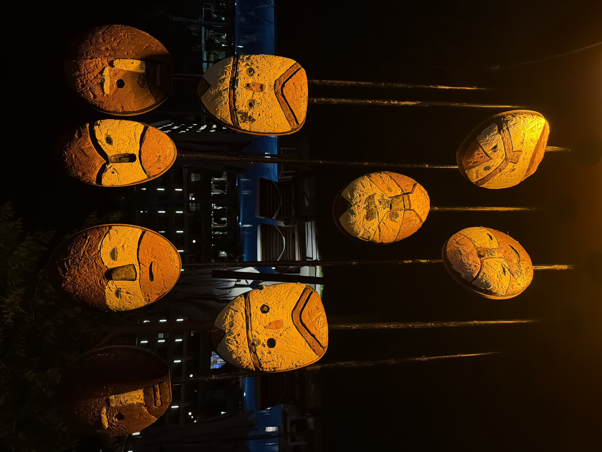 This is a nighttime exterior shot of illuminated masks on poles in Manavgat, Turkey. The masks are of various sizes and feature simplified, stylized faces painted in warm, orange and yellow tones. They are mounted on dark poles of varying heights, creating a staggered display. In the background, a swimming pool and lounge chairs are visible, suggesting the location is a resort or hotel. The lighting is predominantly from the masks themselves, casting an orange glow on the surroundings. There are no people or animals visible in the frame.