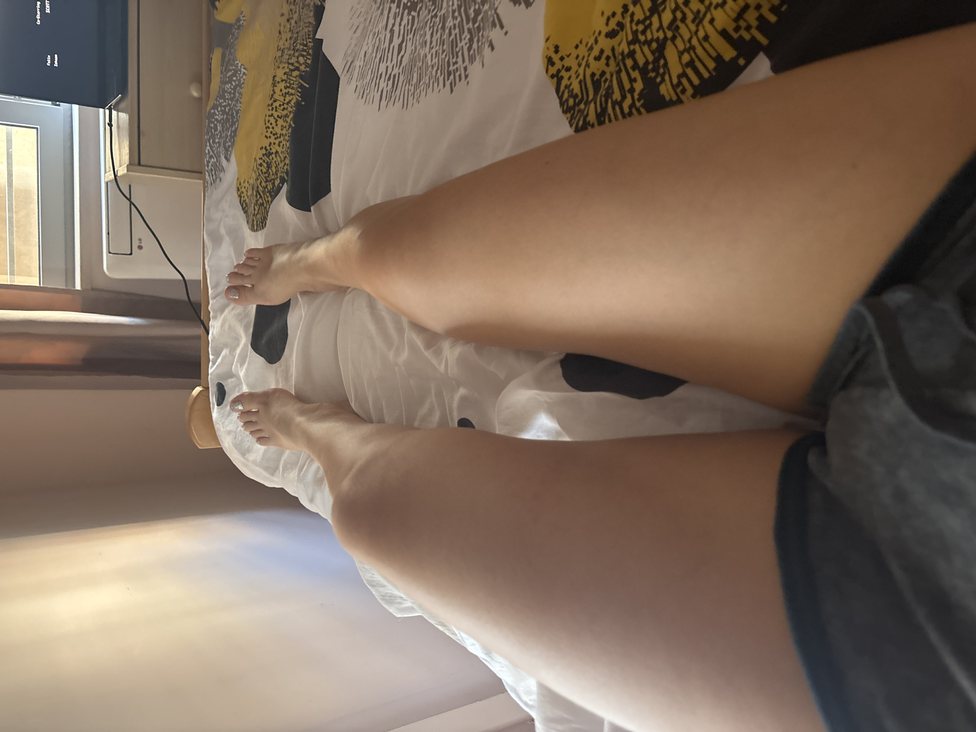 The image shows a person lying on a bed with their legs extended towards the top left corner of the frame. The bed is covered in a patterned white, yellow, and black comforter. The person's legs are fair-skinned and mostly visible from the thigh down. In the background, there is a TV mounted on a wall and a window with daylight filtering in. Based on the presence of a TV, window, and bed, the location appears to be inside a room, possibly a bedroom in a hotel or apartment. However, the city of Budva, Montenegro cannot be confirmed from the image alone.