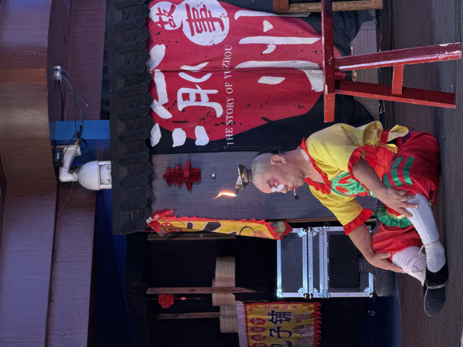 The image depicts a male performer on a stage, likely located in Yuzhong District, Chongqing, China, given the prominent banner text "THE STORY OF YUZHOU." The performer, appearing of East Asian descent, is seated on the dark stage floor with his legs bent and weight resting on his hands. His head is bald on top with dark hair shaved on the sides. He wears theatrical makeup, characterized by a white base with distinct red accents around his eyes. A small, lit oil lamp, emitting a visible flame, is precisely balanced on the crown of his head. He is dressed in a vibrant yellow shirt with red and green patterns, red pants featuring green details, white tights, and black flat shoes. A small microphone is clipped near his mouth.
The stage background on the left consists of a dark wall with stacked wooden steaming baskets, a red hanging lantern, and a yellow banner displaying red Chinese characters. Below these, two silver-grey metallic equipment cases are stacked. The central background features a large red and white banner, styled beneath a black-tiled, traditional Chinese roof-like structure, which proclaims "THE STORY OF YUZHOU" and includes large white Chinese characters. A red paper-cut "喜" (xi, happiness) character is affixed to the wall next to the banner. A red wooden stool is positioned on the stage in the foreground to the right. Overhead, a white dome CCTV camera is mounted on a blue support beam, with visible cables.