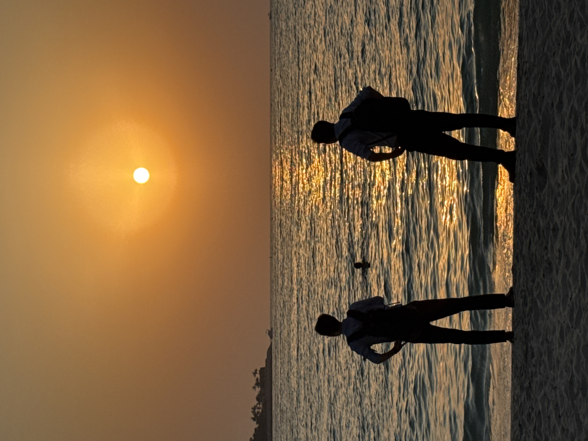 This media file depicts a serene sunset scene at a beach in Dubai, United Arab Emirates. The sun, a bright orb radiating a warm, golden light, hangs low in the hazy sky, casting a shimmering reflection on the calm ocean water. Silhouetted against this backdrop are two individuals, each wearing a backpack and appearing to be enjoying the tranquil ambiance. The first person is positioned closer to the water's edge, their form elongated by the low sun. The second individual stands further back on the sandy shore, their posture suggesting a moment of quiet contemplation or observation. The gentle waves create a textured pattern on the water's surface, with streaks of sunlight breaking through, enhancing the overall peaceful atmosphere. The beach itself appears to be a light-colored sand. The time of day is clearly sunset, indicated by the sun's position and the warm hues of the sky. The weather appears to be clear, with a slight haze contributing to the soft glow of the sunset. The overall mood is one of tranquility and appreciation for nature's beauty. There is no visible text in the media file.