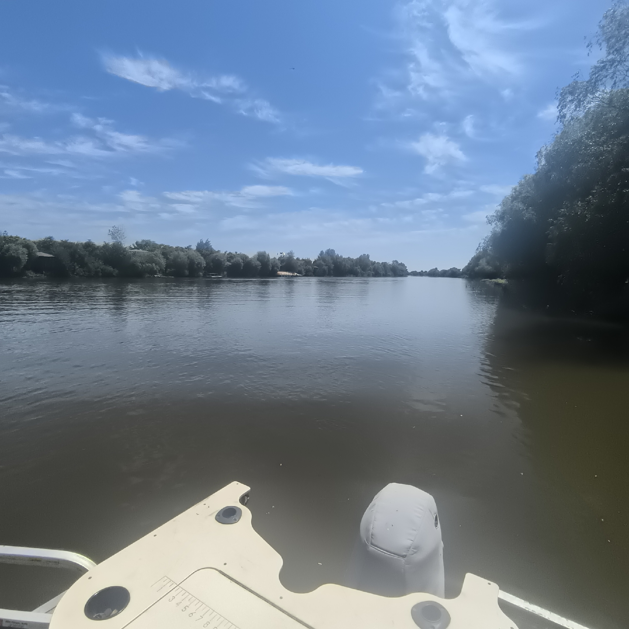 The image shows a body of water, likely a river, under a partly cloudy blue sky. The perspective is from a boat, with the bow and an outboard motor visible in the foreground. The water is calm and reflects the sky. Along the banks of the river, there is dense green foliage, with some structures or houses visible among the trees on the left side. The overall scene suggests a serene, natural environment. The location is specified as Malyy Baklaniy, Russia.