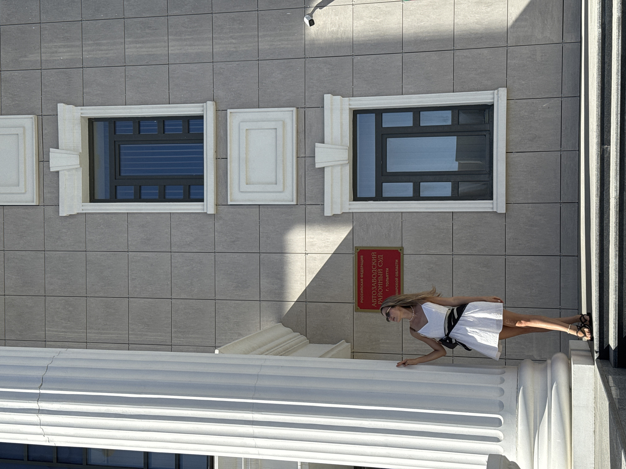 The image depicts a woman in a white dress, black belt, and sunglasses standing near a classical-style building. The building's facade is grey with rectangular tiles. There are decorative architectural elements like window frames and moldings. A sign in Russian is visible, and a security camera is mounted above. The image is taken in Unknown, Unknown.