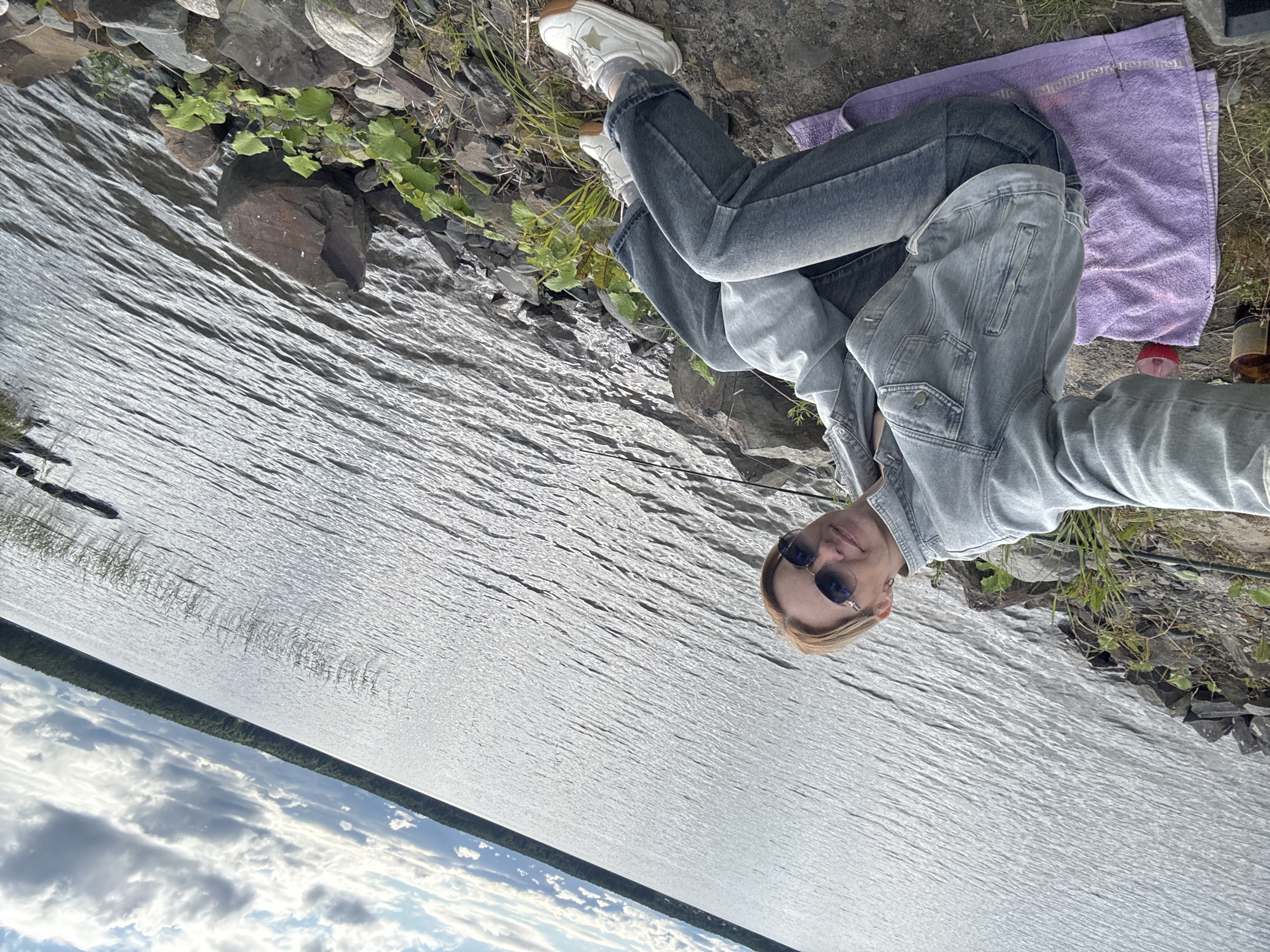 A woman is sitting on the rocky shore of a large body of water, possibly a lake or river, in Shuiskaya Chupa, Russia. She is wearing a denim jacket, jeans, and white sneakers. She is holding up a phone, taking a selfie. A purple towel is spread on the ground next to her, with a bottle and a glass on it. The water is rippling, and the sky is cloudy with patches of sunlight. Trees line the distant shore. The scene appears to be a casual outdoor setting.
