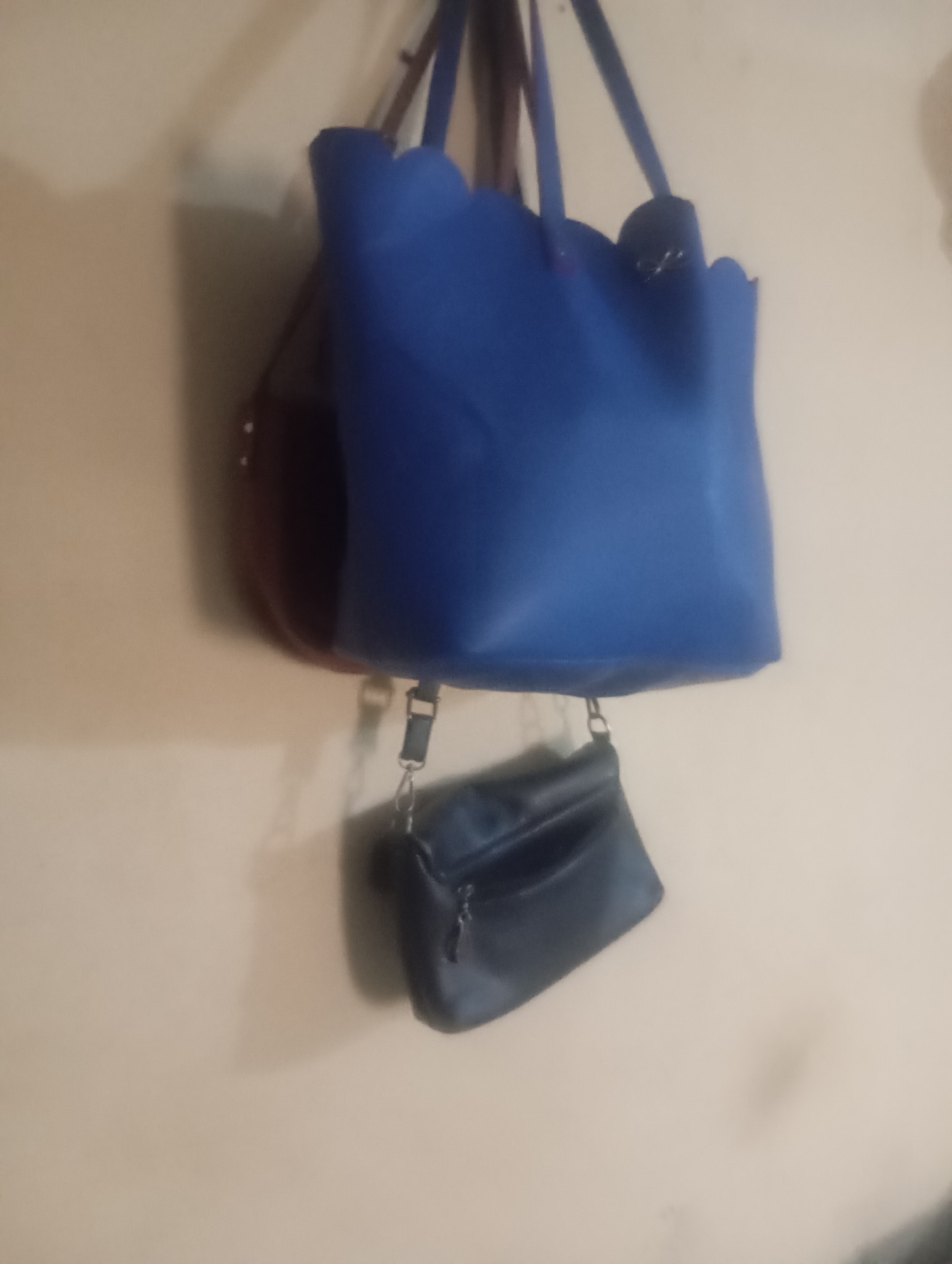 A medium shot photograph, taken from a slightly low angle, captures three handbags hanging on a light-colored wall. The handbags are arranged vertically, with the largest at the top, a medium-sized one behind it, and the smallest at the bottom. The top handbag is a vibrant royal blue with a scalloped edge, its straps made of a darker material. The medium handbag, partially obscured behind the blue one, appears to be a deep reddish-brown. The smallest handbag, hanging below the blue one, is a dark navy blue and has a zipper closure. All handbags appear to be made of synthetic material. The wall has a subtle texture and a shadow is cast from the handbags onto its surface. The image quality is somewhat grainy, suggesting it was taken with a lower-resolution camera or in low light conditions. The scene is devoid of any human presence or activity. There is no indication of the specific location within Jos, Nigeria, beyond being an indoor setting with a wall as the background.