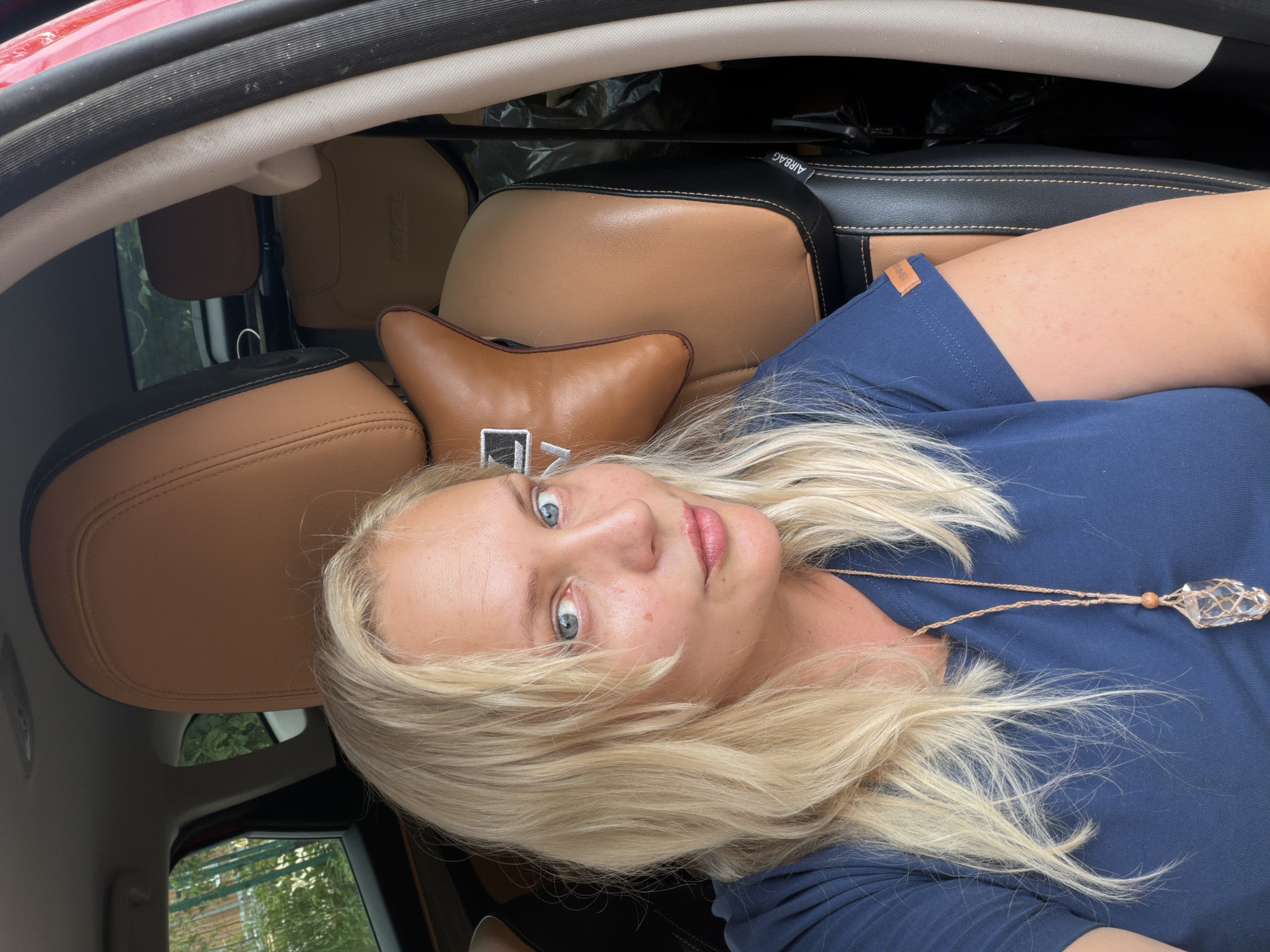 A young woman with blonde hair and blue eyes is sitting in the driver's seat of a car. She is wearing a blue t-shirt and a necklace with a large crystal pendant. The car has leather seats and the windows are tinted. The background is blurry, but it appears to be a residential area. The woman is looking at the camera and smiling. The image was taken in Kazan, Russia.
