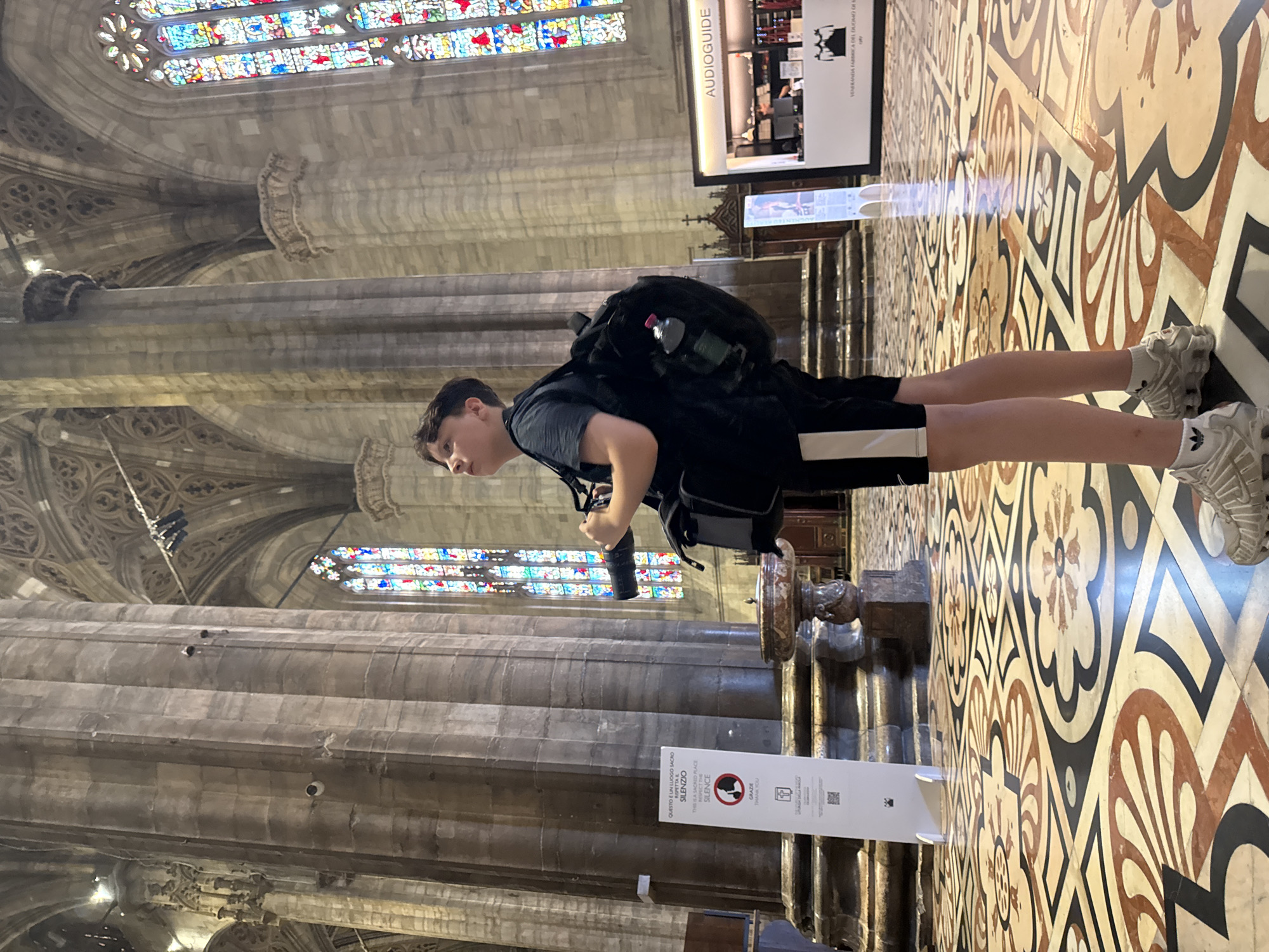 A person stands in a grand cathedral, likely the Duomo of Milan, Italy, given the ornate Gothic architecture and intricate floor tiling. The individual, dressed in black shorts with a white stripe and a dark t-shirt, wears a backpack and holds a camera, appearing to take a photograph or observe the surroundings. The interior features massive stone columns, stained-glass windows casting colorful light, and a highly decorative tiled floor. A sign indicating "AUDIOguide" is visible to the right. The overall scene captures a tourist exploring a significant historical and architectural landmark.