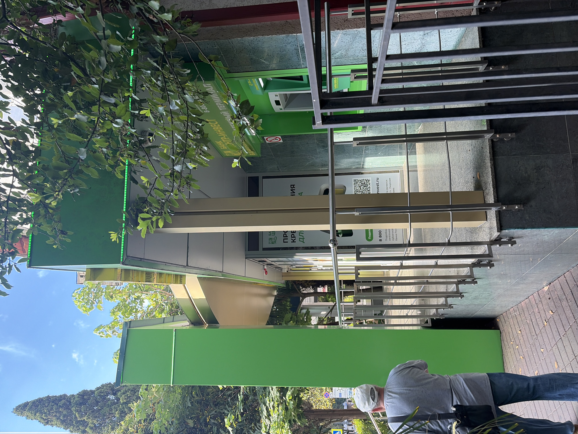 A man wearing a light-colored baseball cap, a gray t-shirt, and blue jeans stands facing away from the camera. He is holding a dark-colored bag with a strap over his left shoulder. To his right, a bright green pillar stands next to a building with green and gold accents. A railing with vertical metal bars separates the sidewalk from a raised area in front of the building. A green ATM is visible inside an alcove of the building. The building has green and marble-like siding, and a sign above the ATM reads "БАНК ЦЕНТР-ИНВЕСТ" (BANK CENTR-INVEST) and "ЭКСПРЕСС ТЕРМИНАЛ" (EXPRESS TERMINAL). Another sign inside the alcove displays a QR code and text. Trees and foliage are present in the background, and the sky is blue with scattered clouds. The ground is paved with bricks. The scene appears to be on a street in Sochi, Russia, as indicated by street signs and architectural style.
