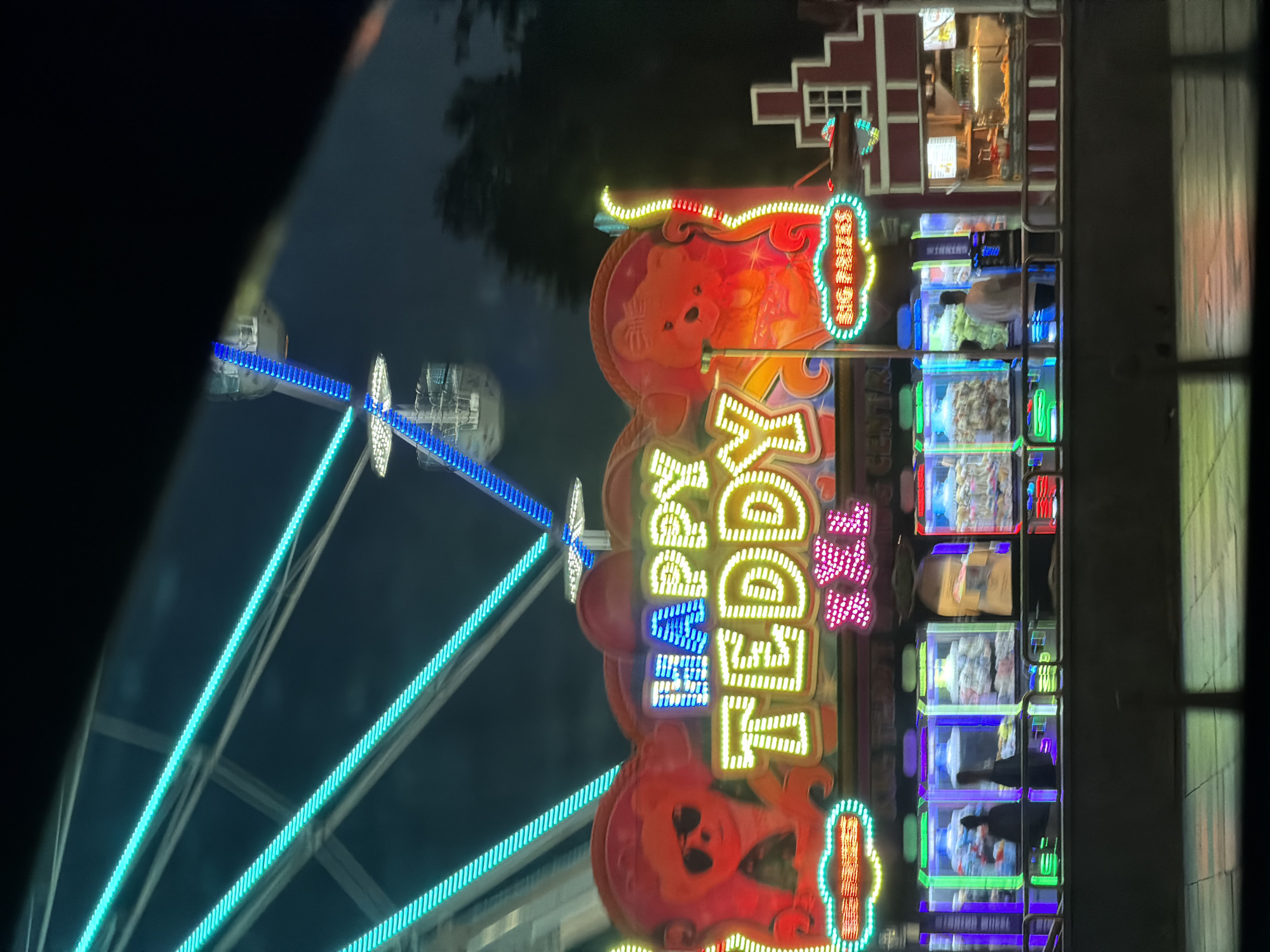 The image depicts a fairground at night. The Ferris wheel, illuminated with blue and white lights, is visible on the left. Next to it is a colorful 'Happy Teddy' sign with teddy bear images. To the right of the sign are illuminated prize displays. Above them is a structure resembling a building facade. The scene is taken in Sofia, Bulgaria.