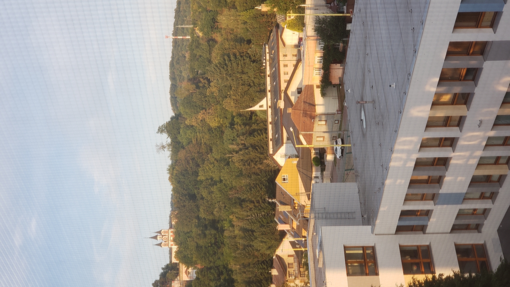 The image captures a suburban residential area with a backdrop of a densely wooded hill. In the foreground, a modern, multi-story building with a light-colored facade and numerous windows is visible. The building's roof is flat and equipped with various vents and pipes. Several other residential buildings with pitched roofs in varying shades of brown and yellow are situated behind it. Some of these houses have visible chimneys and solar panels. A white car is parked on the street between the houses.
Further in the background, a prominent green, tree-covered hill dominates the skyline. Near the top of the hill, a distinct building with multiple towers and a red and white facade, resembling a church or a historic monument, is visible. Another smaller structure, possibly a church with a pointed roof, is nestled among the trees at a lower elevation on the hill. A red and white communications tower stands on the crest of the hill. The sky is a pale blue with some wispy clouds, suggesting it is daytime, likely late afternoon or early morning given the long shadows cast on the buildings.
The image appears to be taken from a higher vantage point, looking across the residential area towards the hill. The presence of buildings with similar architectural styles and the dense vegetation on the hill are characteristic of a European suburban or semi-rural landscape. This scene is consistent with locations within the city of Prešov, Slovakia, particularly areas that transition from urban development to natural hillsides. The style of residential architecture and the presence of a prominent landmark on the hill further align with observations of the Prešov area.