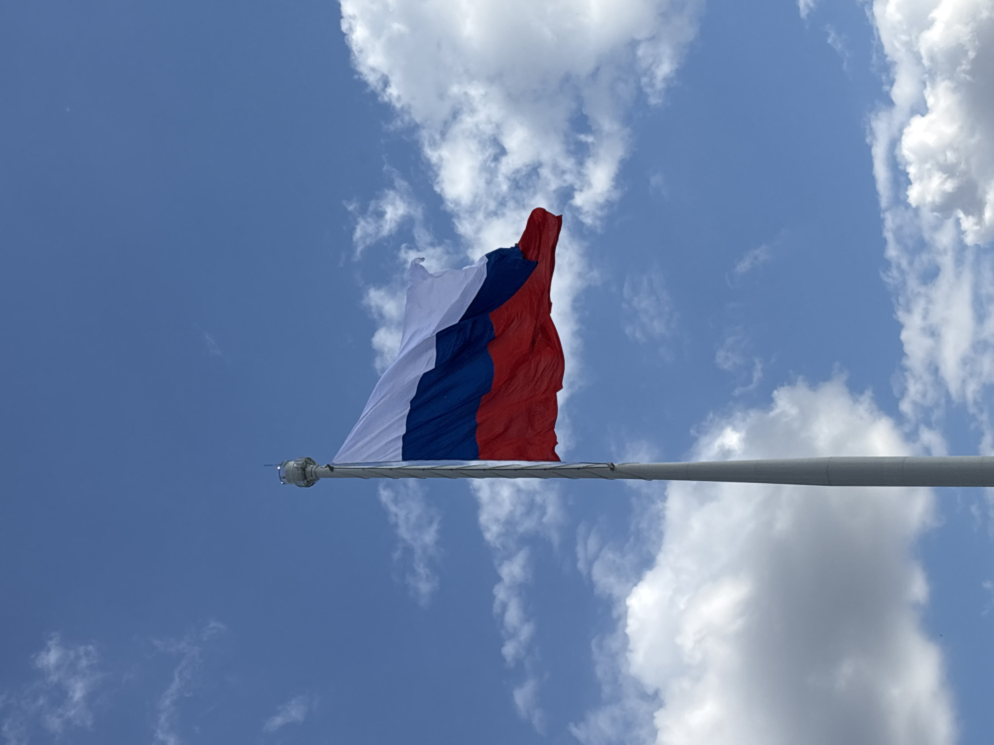 A low-angle shot shows the Russian flag waving against a blue sky with scattered white clouds. The flag, composed of three horizontal stripes of white, blue, and red, is attached to a tall, gray flagpole that extends upwards from the bottom of the frame. The top of the flagpole features some equipment, possibly for lighting or weather monitoring. The scene is likely outdoors, with the flag serving as a prominent visual element against the expansive sky. The image is a clear and straightforward depiction of a national symbol in a natural setting.