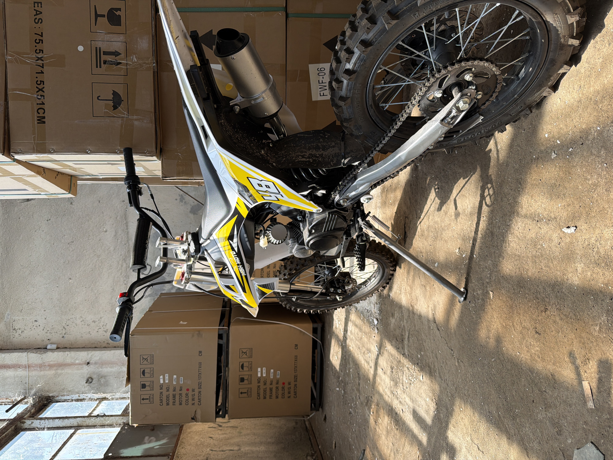 An off-road motorcycle is parked on a concrete floor in what appears to be a warehouse or storage unit. The motorcycle is predominantly white with yellow and black accents, featuring the number 48 prominently on its side. It has knobby tires suitable for off-road use and a silver exhaust pipe. The background consists of stacked cardboard boxes, some with shipping labels and hazard symbols. Natural light streams in from a large, grimy window on the left side of the frame, casting shadows across the floor. The overall environment suggests a commercial storage space, possibly for new inventory.