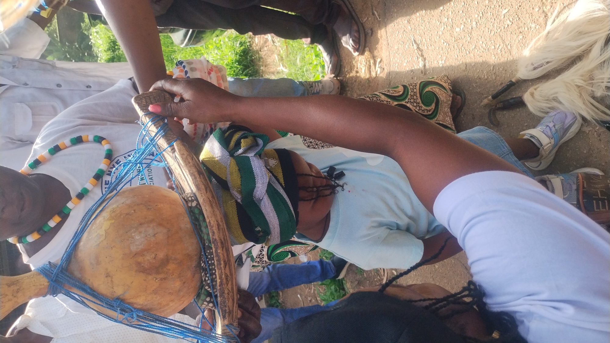 In Jos, Nigeria, at an outdoor event, a woman holds a gourd tied with blue string above a young child wearing a light blue t-shirt and a colorful headwrap. The child looks down, with several dark beads attached to their braided hair. In the background, other individuals are visible. One person, wearing a white shirt and a colorful beaded necklace, stands behind the gourd. Another person, dressed in a white shirt and a grey shirt underneath, is partly obscured. To the right, a person in dark pants and sandals stands with their legs visible. The ground is paved with asphalt, and the scene is illuminated by natural daylight. Two white, feather-like objects with dark handles lie on the ground in the foreground.