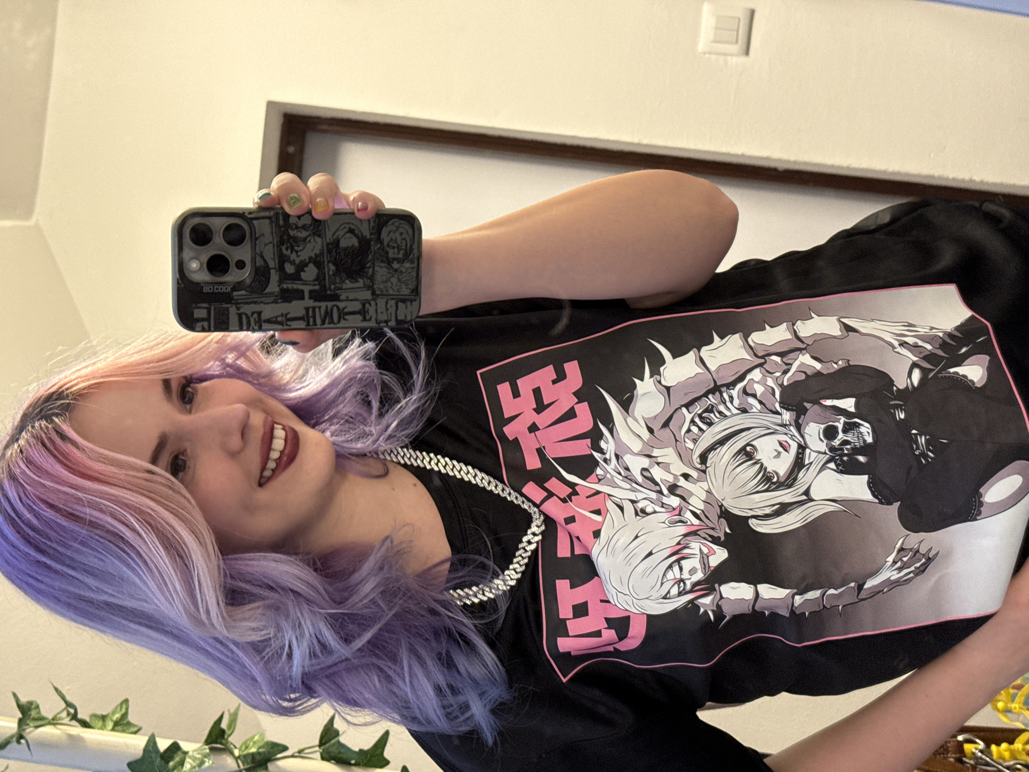 A woman is taking a selfie in front of a mirror. She has light skin, pink and purple hair, and is smiling. She is wearing a black t-shirt with a large graphic print of anime-style characters with skeletal elements, and a thick silver chain necklace. She is holding a smartphone with a patterned case in her left hand, obscuring part of her face.
The background consists of a white wall with a light switch and a picture frame. An artificial ivy garland is visible in the lower left corner of the image, possibly adorning the mirror or a nearby surface.
Based on this information, it is impossible to determine the location of the photograph to be within the city of City of Zagreb, Croatia.
