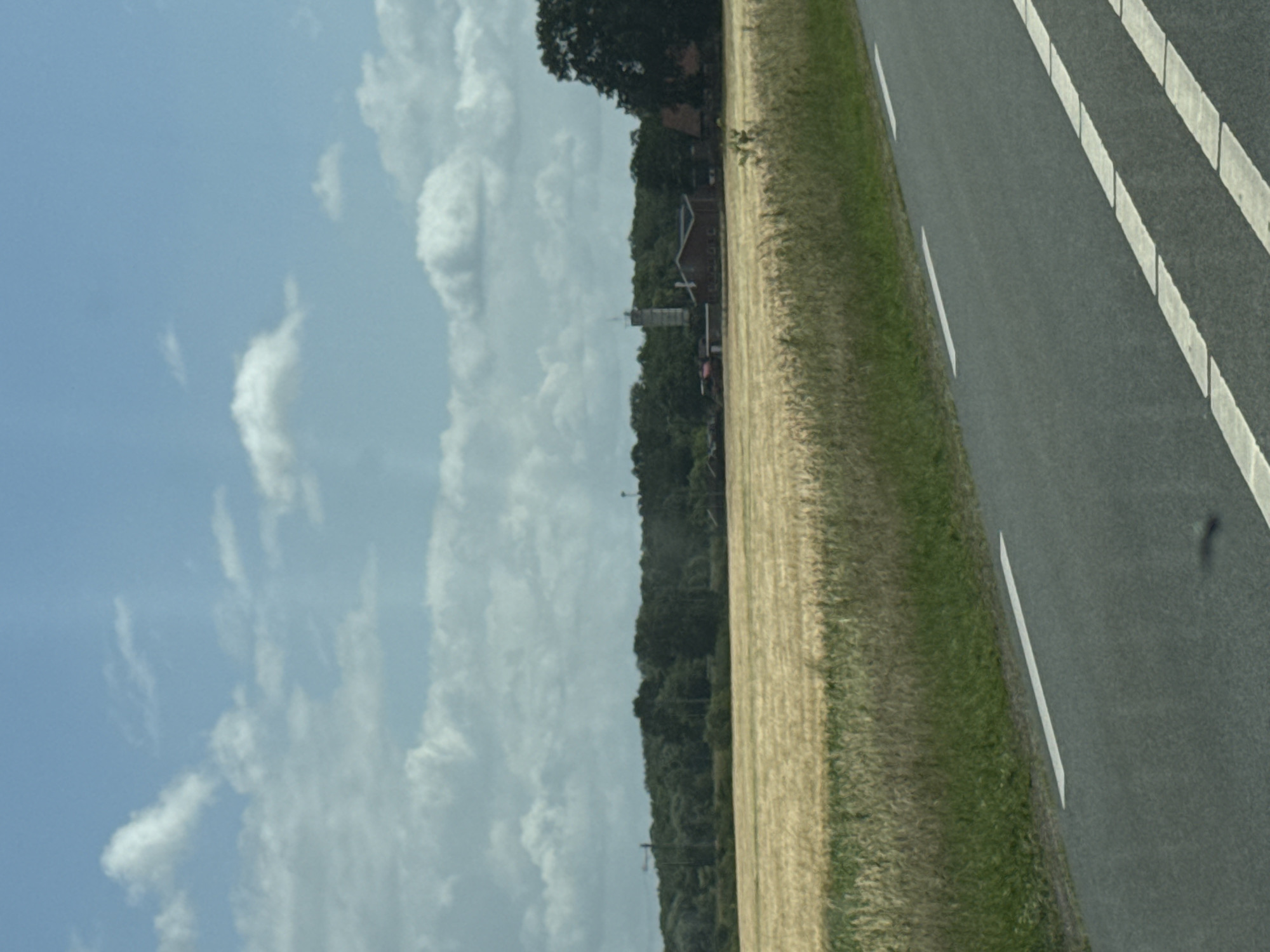 The image depicts a rural landscape with a road in the foreground. A field of dry, golden crop, likely wheat or barley, stretches across the middle ground. Behind the field, a dense line of green trees forms a natural boundary. A cluster of buildings, possibly a farm or small industrial complex, is visible amongst the trees, with a tall, slender tower structure also present. The sky is a pale blue, dotted with scattered white clouds. The road in the foreground has white lane markings. The scene is static and appears to be a daytime photograph taken from a moving vehicle.