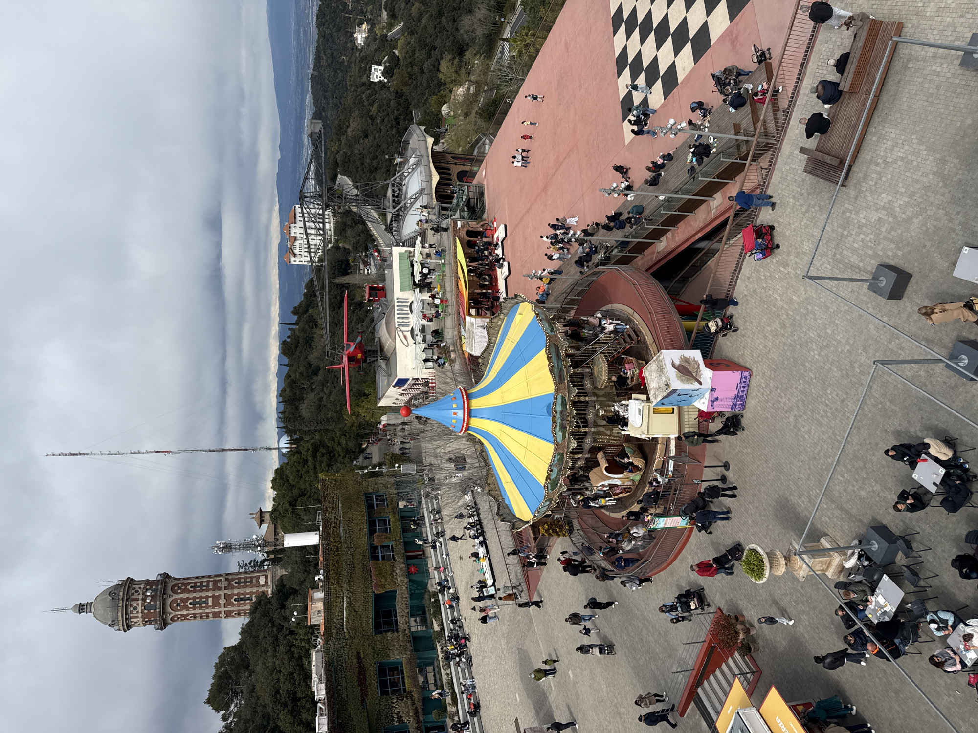 The image shows a high-angle view of an amusement park in Barcelona, Spain. The scene includes a carousel with a blue and yellow striped top, a checkered platform, and several structures. Many people are scattered throughout the park. The Torre de Collserola is visible in the background, along with forested hills and a cloudy sky. Several benches and outdoor tables are also scattered throughout the park.