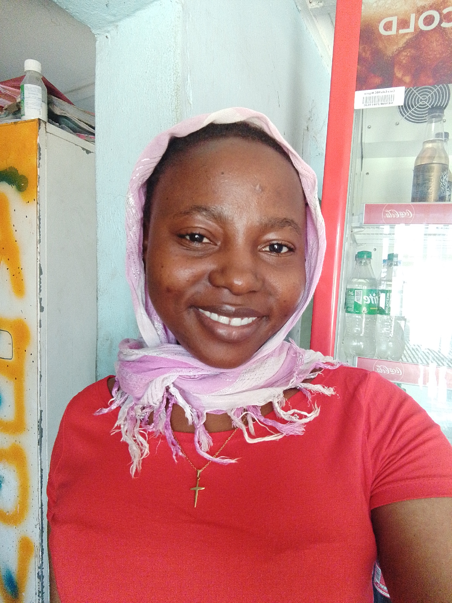 A medium shot depicts a Black woman with smooth, brown skin, wearing a pink and white scarf on her head and a red t-shirt. She has a gold cross necklace around her neck and is smiling. The background includes a blue-green wall, a yellow and white cabinet with colorful graffiti, and a red Coca-Cola cooler. The location is in the city of Unknown, Unknown.