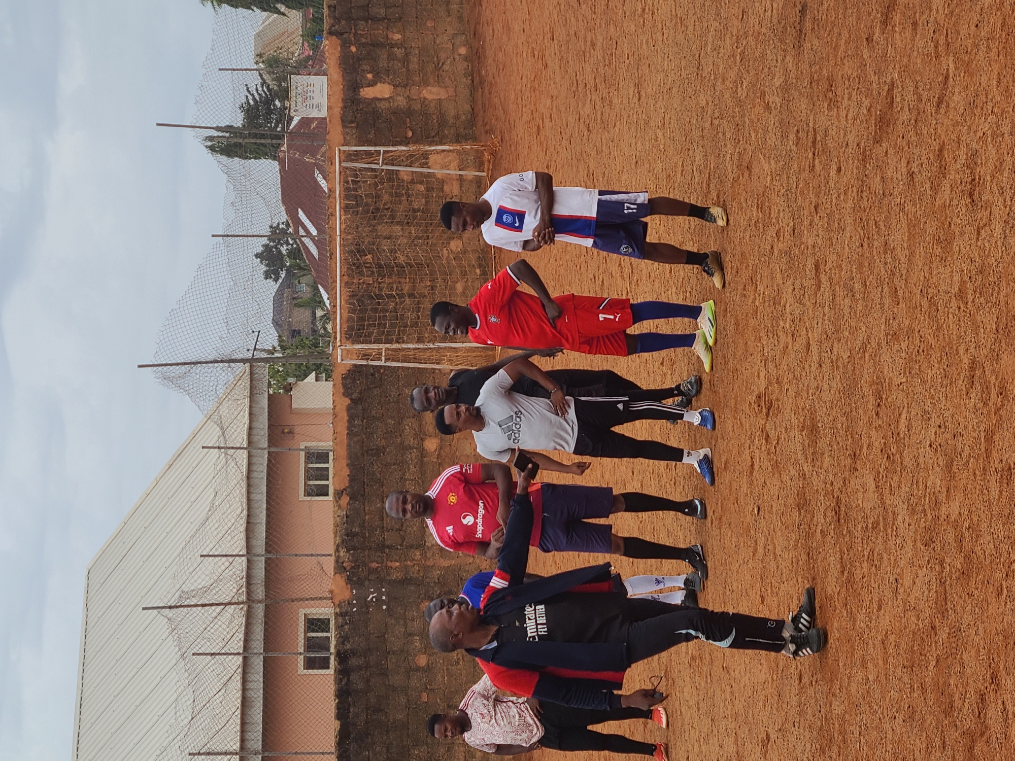 A group of people are standing in a line on a reddish-brown dirt field in Unknown, Unknown. There are buildings and fences in the background, along with a soccer net. The people are wearing various types of athletic clothing.