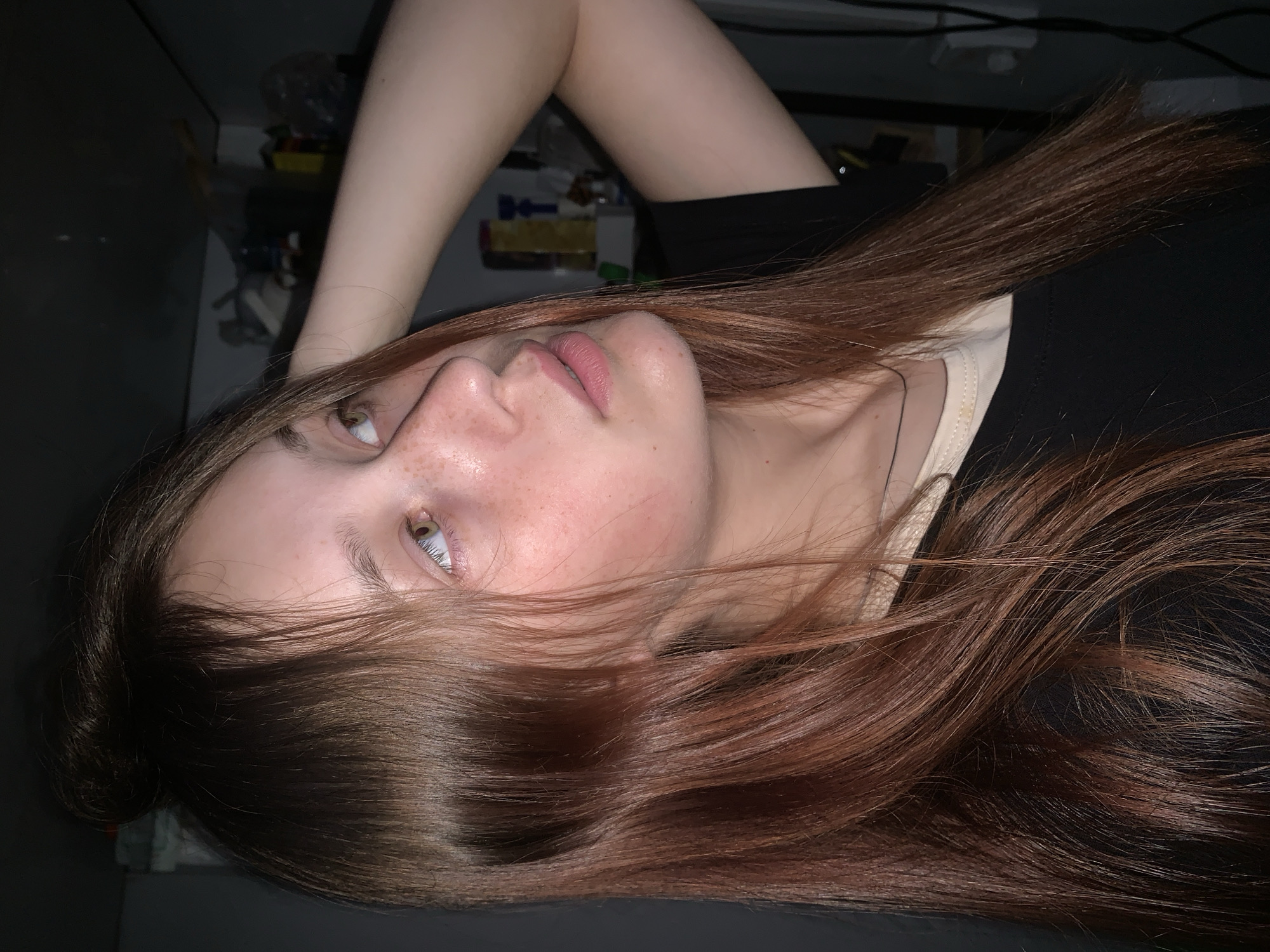 A young person, likely female, is depicted in a medium close-up, rotated 90 degrees clockwise, against a dimly lit indoor background, possibly in Фоки, Russia. She lies in a relaxed pose, with her head resting on a dark surface. Her gaze is directed upwards and slightly to the right of the frame, revealing striking light-colored eyes, possibly green or blue, with prominent pupils. Freckles are visible across her nose and cheeks. Her lips are slightly parted, exhibiting a soft pink color.
Long, reddish-brown hair, appearing lustrous and healthy, flows around her head and across her shoulders, with some strands delicately framing her face. She is wearing a black top, with a cream or light-colored garment visible underneath at the neckline. One bare arm is visible, bent at the elbow and positioned above her head.
The background is dark and largely out of focus, suggesting low light conditions, indicative of evening or night. Vague, indistinct shapes of objects, possibly including bottles, boxes, and cables on shelves or surfaces, can be faintly discerned in the upper left portion of the image. The setting appears to be a casual indoor space, possibly a bedroom. The subject's expression is calm and contemplative. There is no visible text in the image.