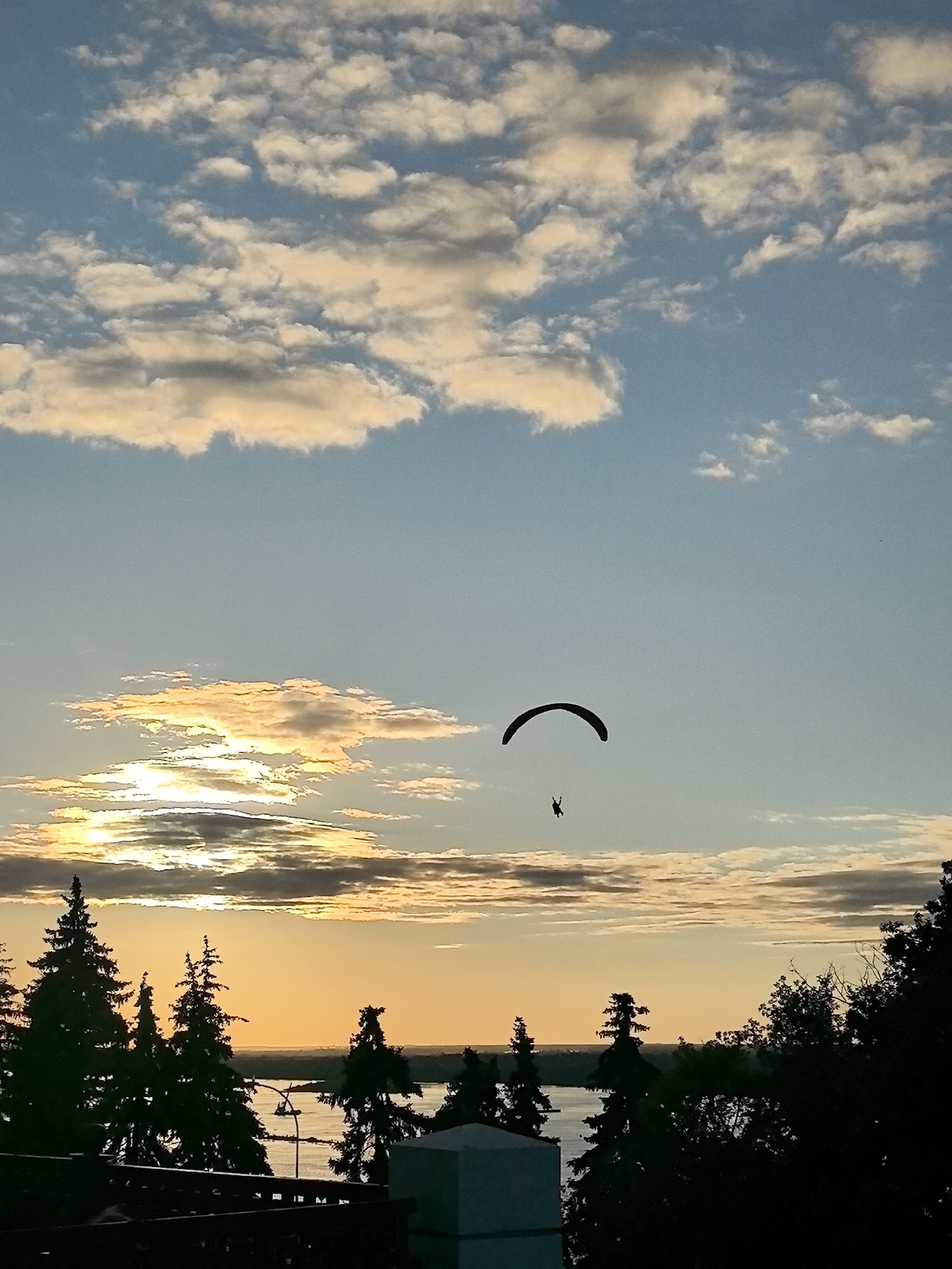 The image depicts a paraglider in silhouette against a sky with scattered clouds during sunset. The sky transitions from a light blue at the top to a warm orange glow near the horizon, where the sun is setting behind a line of clouds. A paraglider is visible in the upper middle portion of the frame, suspended in the air. In the foreground, silhouetted evergreen trees line the bottom of the image, with a body of water visible beyond them. A dark railing and a white structure are also visible in the foreground. The setting suggests an outdoor recreational area overlooking a natural landscape, possibly a park or a waterfront in the city of Nizhny Novgorod, Russia.