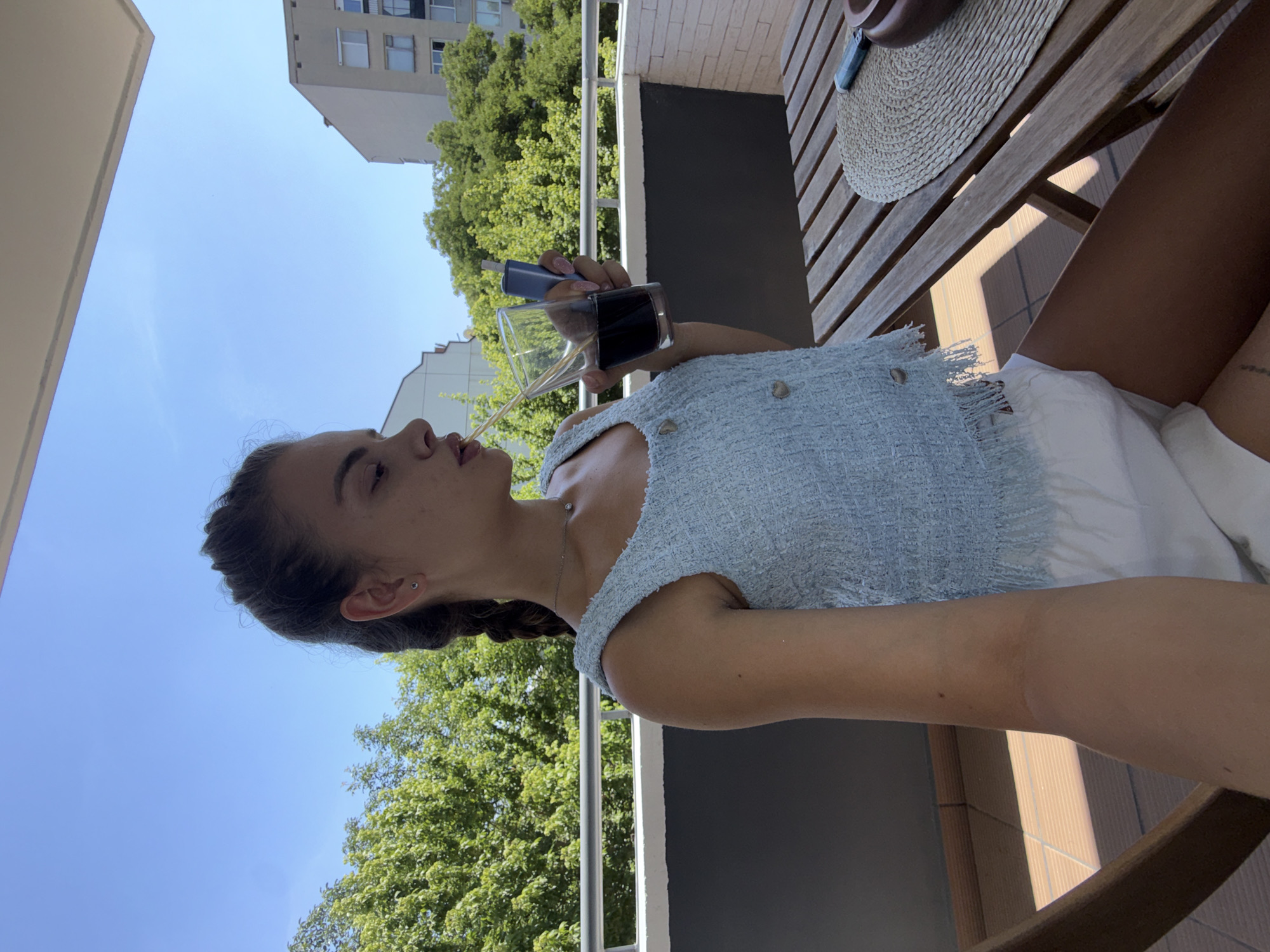 The image captures a young woman sitting on a balcony with a clear blue sky and trees in the background. She is wearing a light blue, textured sleeveless top with buttons and white shorts. The woman has her hair in a braid and is drinking from a glass with a straw, holding a blue electronic device in her other hand. A wooden table with a straw placemat is visible in the foreground. The building visible in the background suggests an urban environment. Based on the architectural style and common building designs in Plovdiv, Bulgaria, this scene is likely situated on a residential balcony in the city.