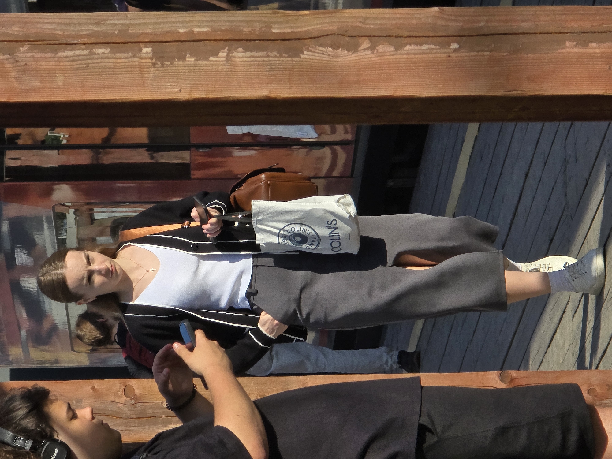 The image shows a woman and a man on a wooden bench, possibly in Khimki, Russia. The woman is wearing a white tank top, a black cardigan with white stripes, gray trousers, and white sneakers. She has a brown purse and a "Colin's" shopping bag next to her. She is holding a blue smartphone in her hand. The man is lying on the bench with headphones on. He is wearing a black t-shirt. The background consists of wooden structures and reflections. The orientation of the image is rotated.