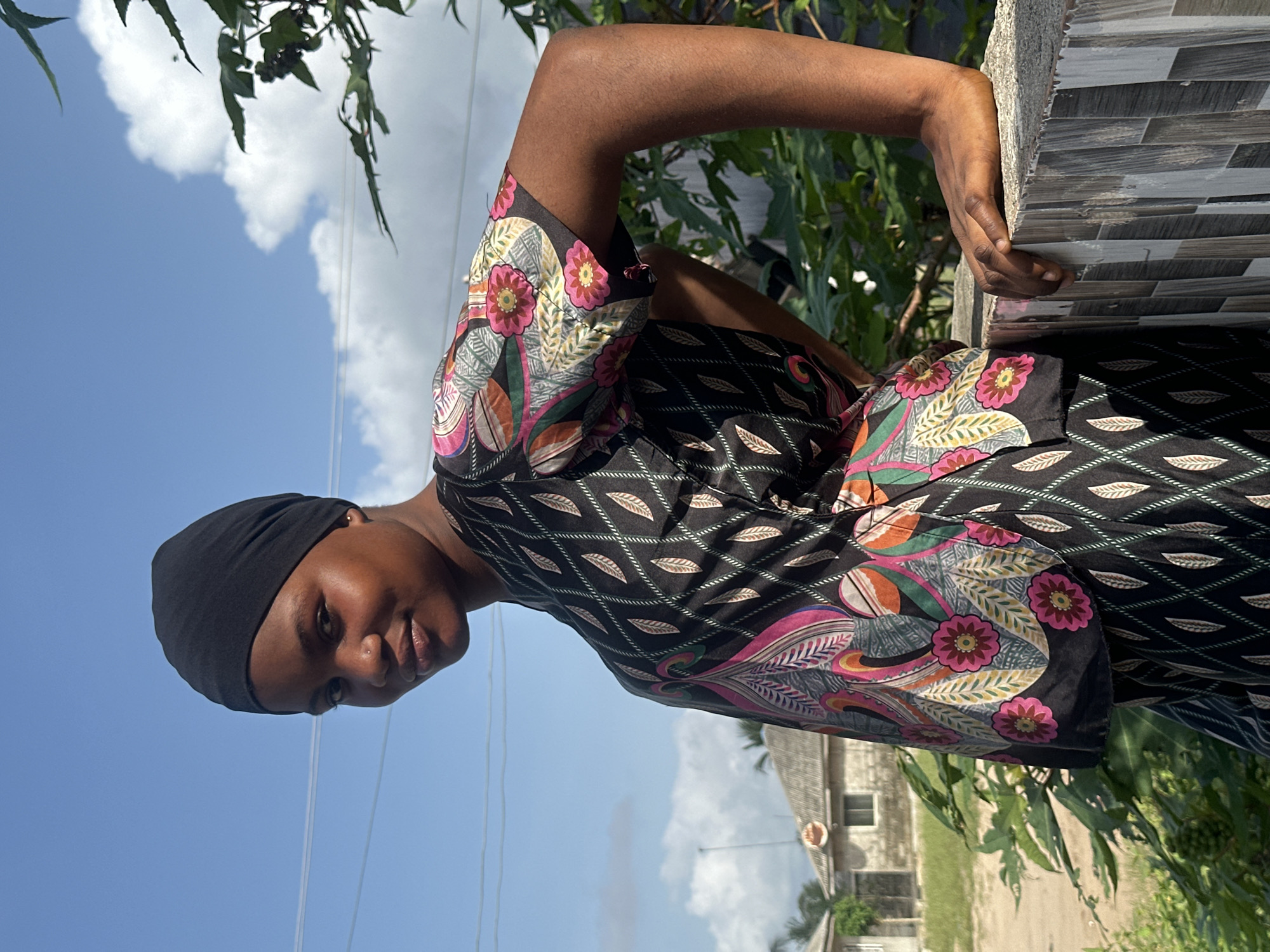 A young Black individual, likely female, is depicted in a sunlit outdoor portrait, situated in Badagry, Nigeria. She wears a plain black head covering and a visually striking black shirt featuring intricate floral and leaf patterns in vibrant hues of pink, green, orange, and white. Her dark-skinned arm is bent at the elbow, with her hand resting on a textured grey and white tiled wall or ledge, which displays a rectangular, brick-like pattern. She looks towards the viewer with a neutral and composed expression.
The background is dominated by a clear, bright blue sky with scattered white cumulus clouds, indicating sunny weather, likely around midday or early afternoon. Lush green foliage, consistent with trees or bushes, frames the scene, appearing both in front of and behind the subject. In the distance, faint, light-colored buildings with dark roofs are visible, suggesting a residential or semi-urban environment. Thin, dark lines, possibly power lines, cross the sky. The image captures a serene moment within a natural and built environment in Nigeria. There is no visible text.