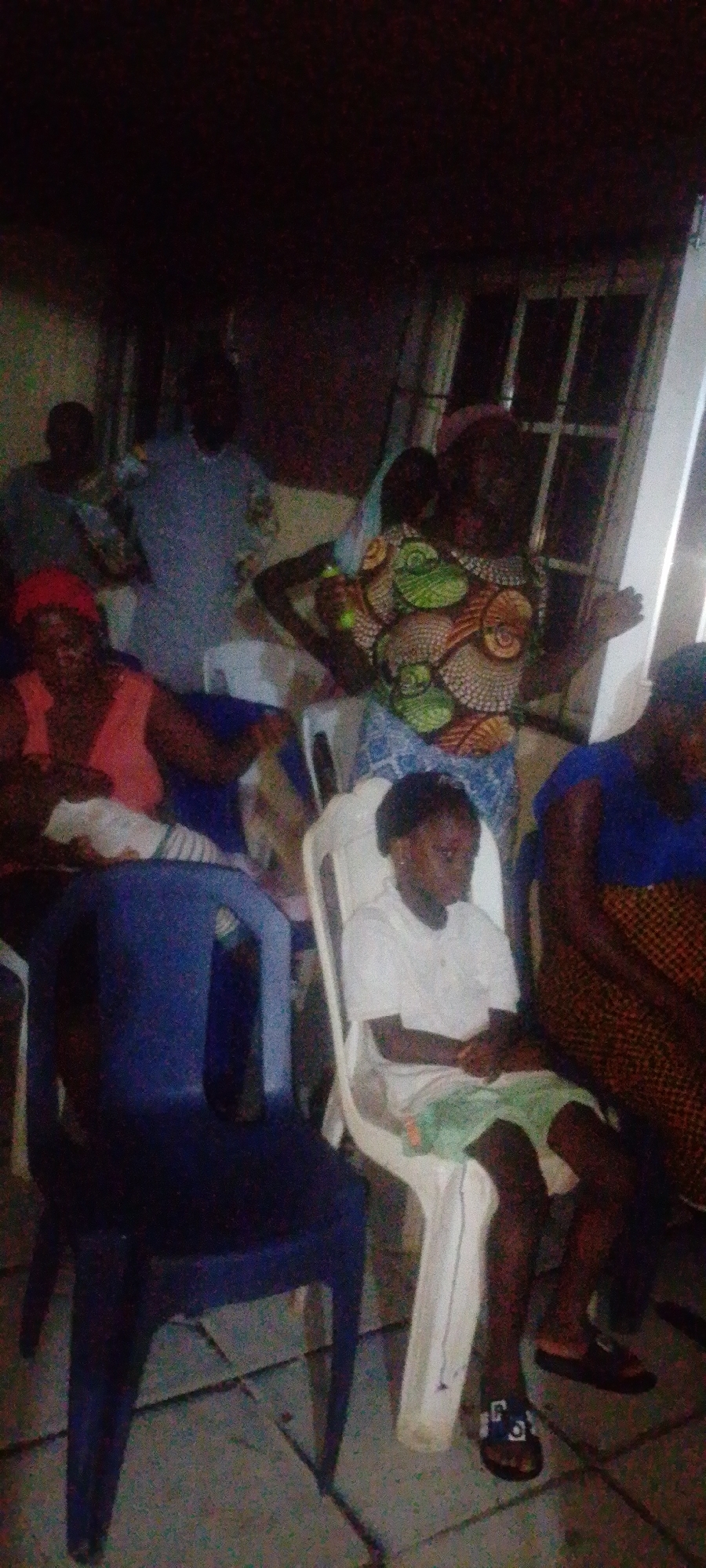 The image shows a group of people gathered indoors, likely a home or community space, in Lokoja, Nigeria. The lighting is dim, and the focus appears to be on those in the foreground. Several people are seated on plastic chairs, with a blue chair prominently placed in the left foreground. A young girl is seated on a white chair, wearing a white shirt and green shorts, her arms crossed. An older woman stands behind her, wearing a colorful patterned top and a blue skirt. Other individuals are seated and standing, but details are less clear due to the lighting and focus. A window with multiple panes is visible in the background. The environment suggests a casual gathering.