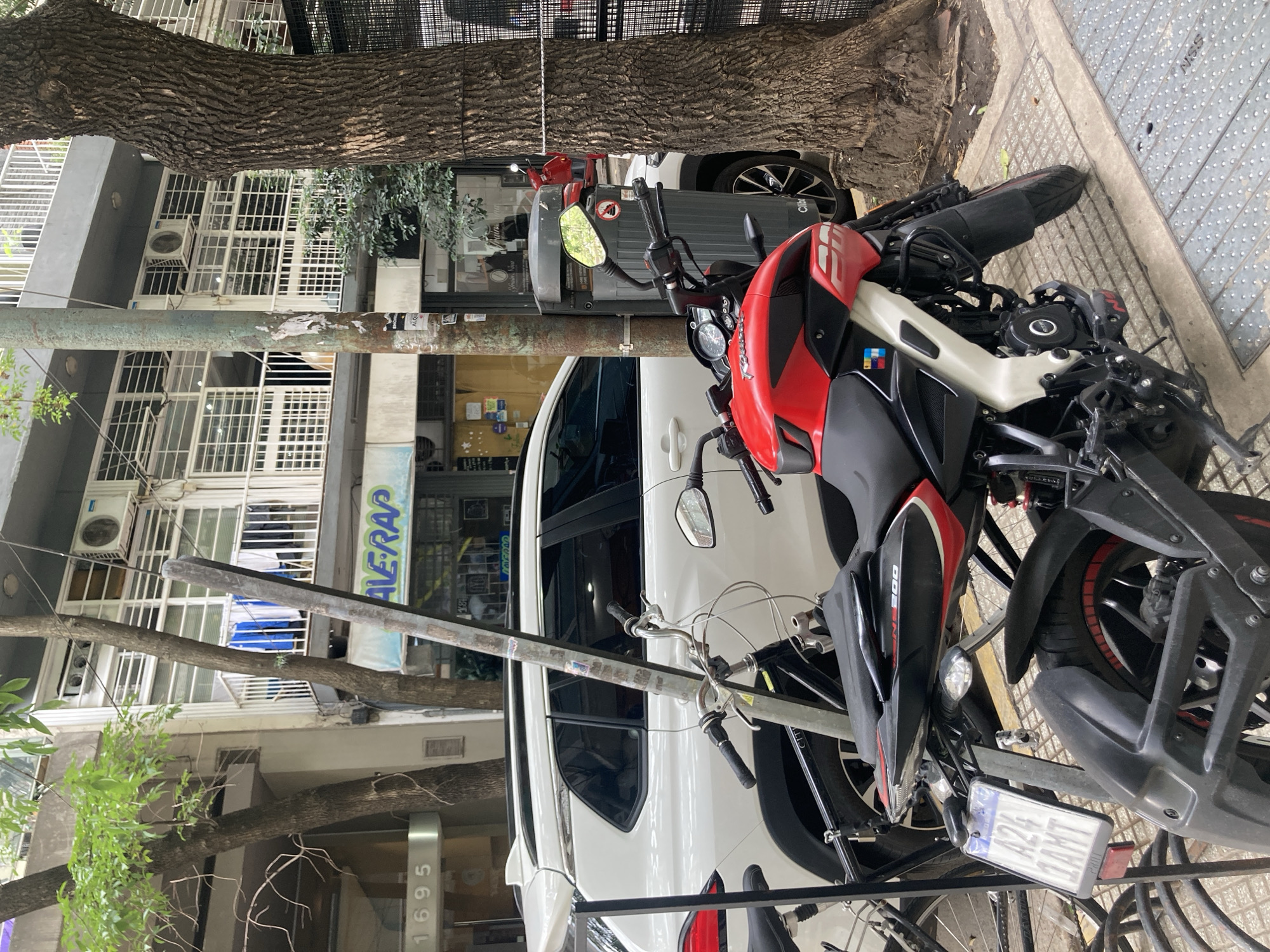 The image depicts a street scene with a red and black motorcycle parked on the sidewalk. It is secured to a metal bicycle rack. A white car is parked behind the motorcycle. In the background, buildings with balconies and shops are visible. A tree trunk is also present in the foreground and background. The scene appears to be a typical urban setting.