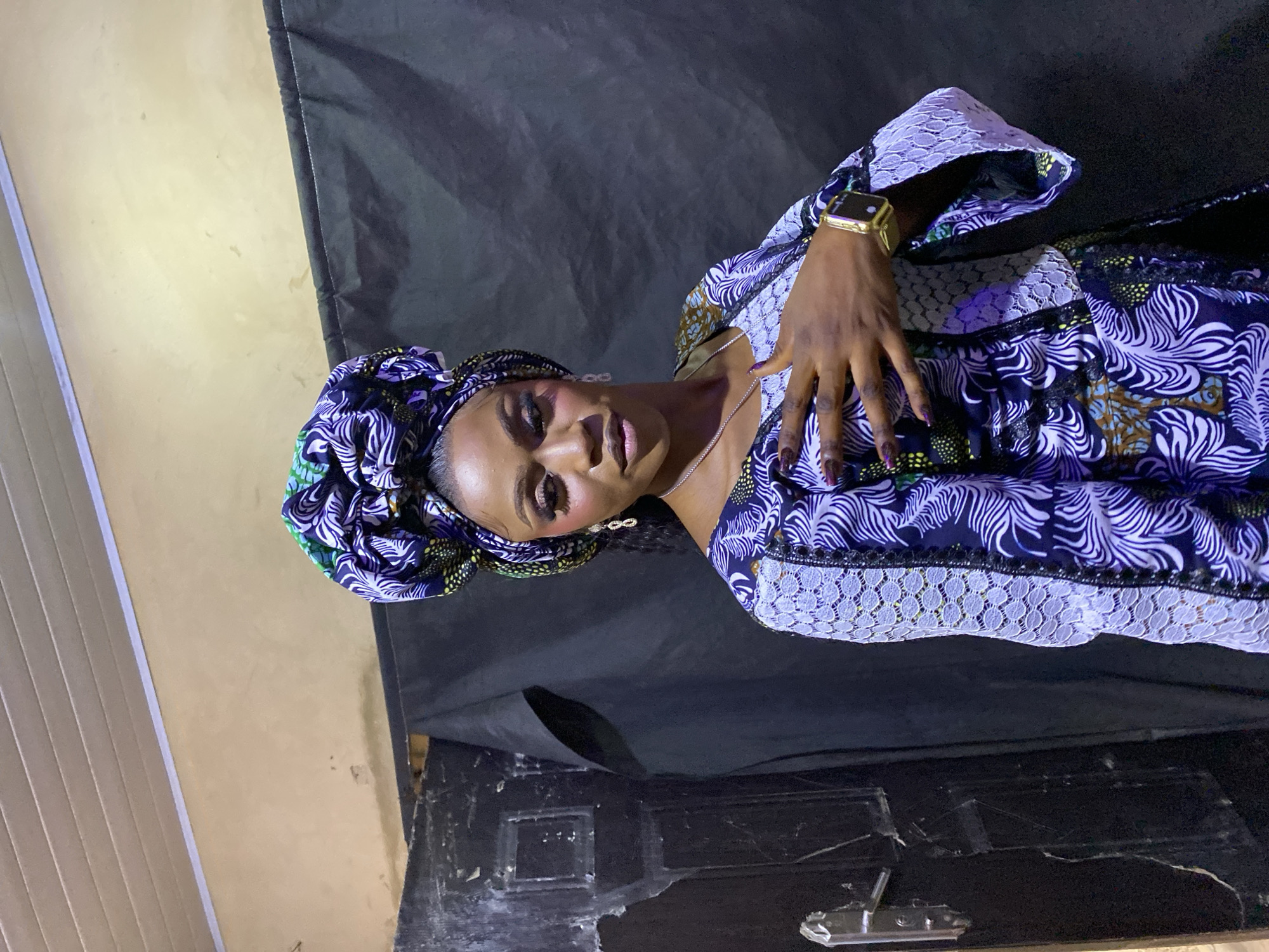 This is a portrait of an African woman, captured indoors against a mixed background. She looks directly at the camera with a subtle, confident smile.
The woman wears a vibrant, patterned garment with a matching headwrap. The fabric is predominantly dark blue/purple with light blue/white leaf-like patterns and accents of yellow and green. White lace detailing is visible on the sleeves and along the garment's edges. She accessorizes with a gold-colored digital wristwatch on her left wrist, a thin silver necklace, and earrings resembling infinity symbols. Her makeup accentuates her eyes and lips, and her fingernails are painted a dark, possibly plum, color.
The background behind her left side consists of a light, possibly beige-painted wall. To her right and behind her body, a dark, textured material, likely a fabric backdrop, is visible. The lower part of the image shows a dark, worn surface, potentially a floor or furniture. A sliver of light-colored vertical blinds or paneling can be seen on the far left.
The woman is posing for the photograph, with her left hand gently resting near her chest, highlighting her watch and manicured nails. The indoor setting and direct lighting suggest artificial illumination. While the image is set in Kuru Dungus, Nigeria, the visual elements within the photo are general to an indoor shoot, though her attire and appearance are culturally consistent with the region. There is no visible text in the image.