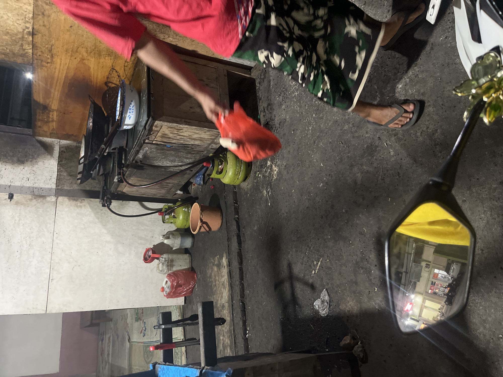 A person wearing a red shirt and camouflage-patterned shorts is standing in a dimly lit outdoor area. They are holding a red cloth in their right hand. In the background, there is a stove with pots and pans, gas cylinders, and various containers. The ground is covered in dirt and debris. A motorcycle side mirror in the foreground reflects a building with several parked motorcycles. The scene appears to be an outdoor food stall or workshop.