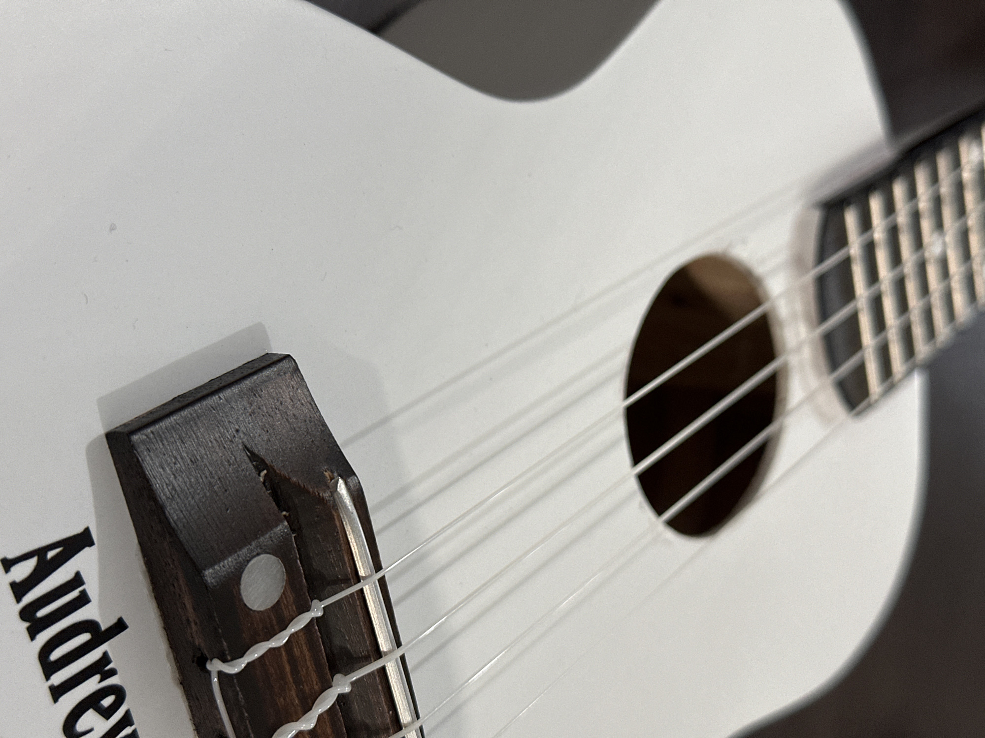 A close-up, eye-level, interior shot captures a white ukulele positioned on a dark, possibly wooden surface. The ukulele is angled diagonally, with its tuning pegs and fretboard visible in the upper left of the frame, blurred by the shallow depth of field. The soundhole is also in the upper portion, with strings extending downwards. The bridge, a dark wooden piece, is in focus in the lower right quadrant, supporting the white strings which are tied with knots. Black text, partially visible and reading "Audrey", is present on the white body of the ukulele in the lower right corner. The image is well-lit, with the light source appearing to be from the upper left, casting subtle shadows on the ukulele's body. The overall impression is one of an object at rest, possibly being displayed or stored, in a dimly lit room. The precise location within Bulakrejo, Indonesia cannot be determined from the visual information provided.