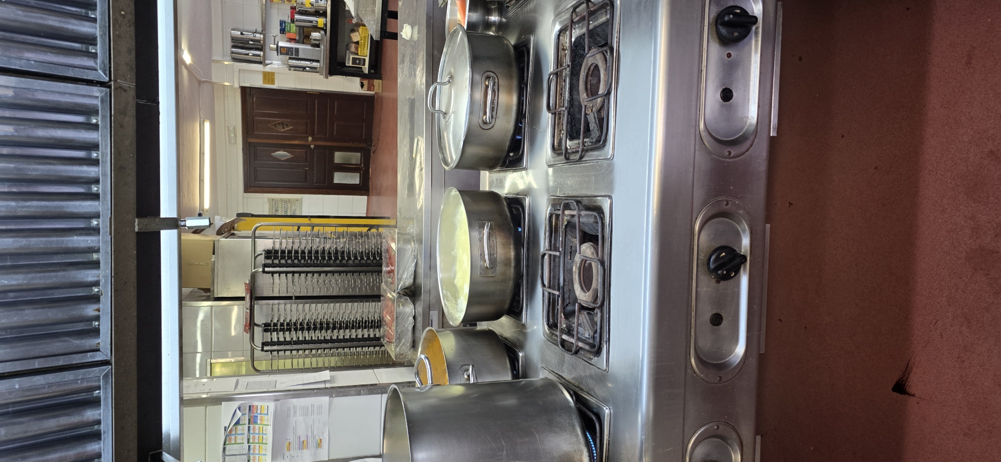 The image shows a commercial kitchen at a restaurant in Sallent de Gállego, Spain. The focus is on a stainless steel stove with four burners, two of which are in use. On the left burner, a large silver pot containing a yellow liquid is cooking. Next to it, on the second burner from the left, is a silver pot with a lid, containing a light yellow liquid that appears to be boiling. To the right of these, two more burners are visible, one with a silver pot with a lid, and the last one appears to be off. The stove has control knobs below the burners. In the background, a stainless steel rack holding trays covered in plastic wrap, containing raw meat, is visible. Further in the background, there are double wooden doors and kitchen equipment. The overall environment is that of a busy, functional kitchen.
