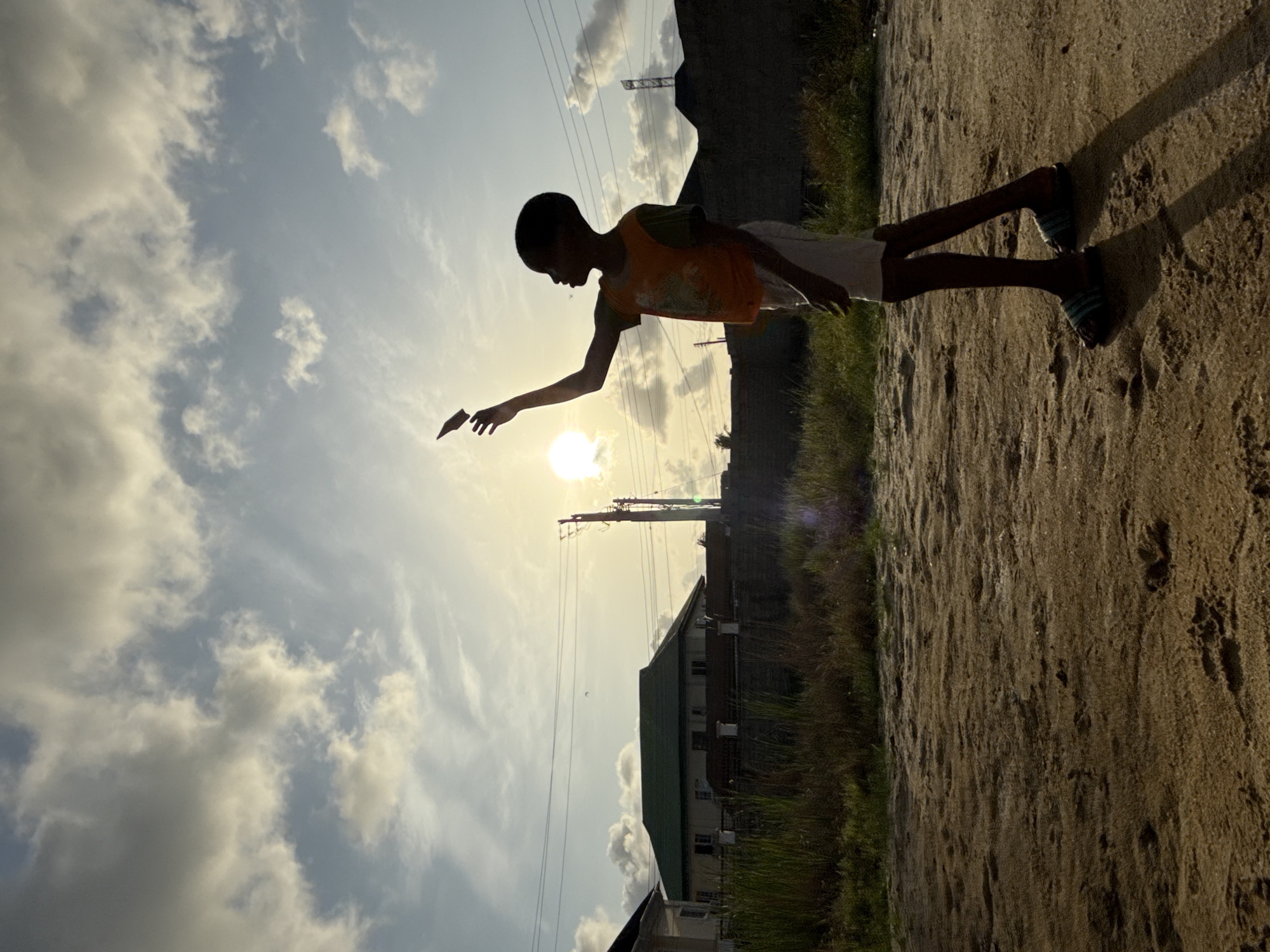 A silhouette image taken outdoors in Ado, Nigeria, shows a young boy standing on a dirt surface with a green grassy edge. He is facing the sun, which is partially obscured by clouds, and he is holding what appears to be a small, flat object in his outstretched hand. The background includes buildings, power lines, and a cloudy sky. The image is taken from a low angle, giving the impression that the boy is reaching for the sun.