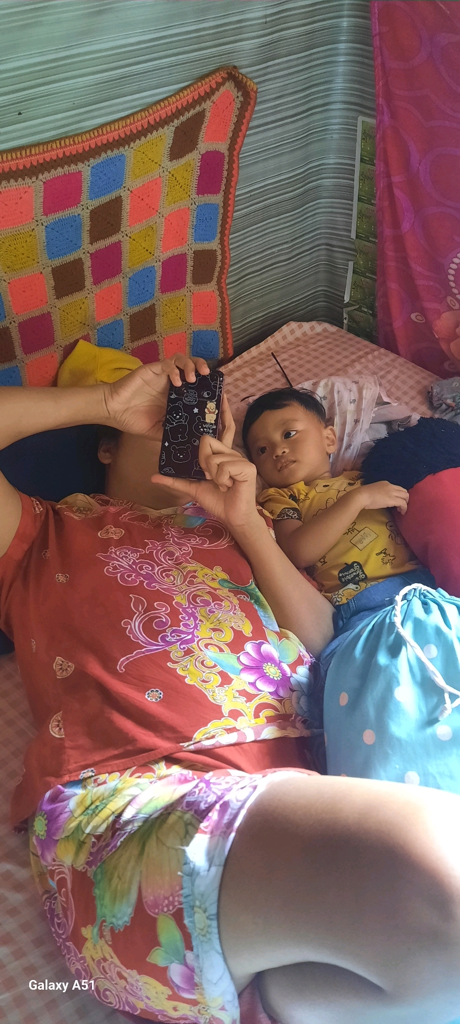 In a bedroom in Ploso, Indonesia, a person is lying on their side, holding a dark-colored smartphone with a Winnie the Pooh case. A young child is lying next to them, looking towards the phone. The person is wearing a red t-shirt with a floral pattern and colorful shorts. The child is wearing a yellow t-shirt with a graphic print and blue shorts. A light blue drawstring bag with white polka dots is positioned next to the child. The room's background features a striped wall and a colorful crocheted blanket.